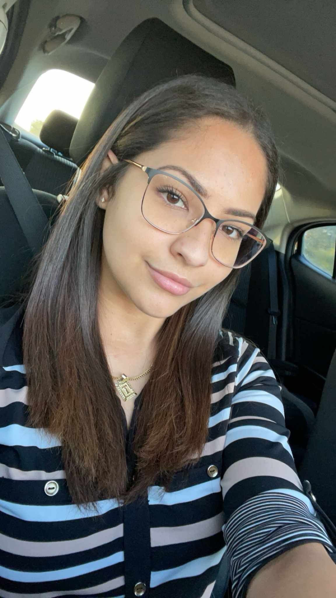 Woman in car, wearing glasses, striped shirt, smiling at camera.