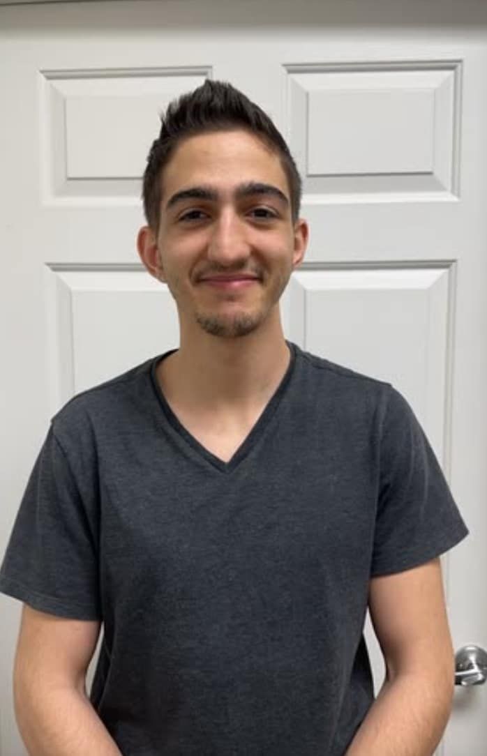 Man with short dark hair and stubble smiles, wearing a gray V-neck shirt, stands in front of a white door.
