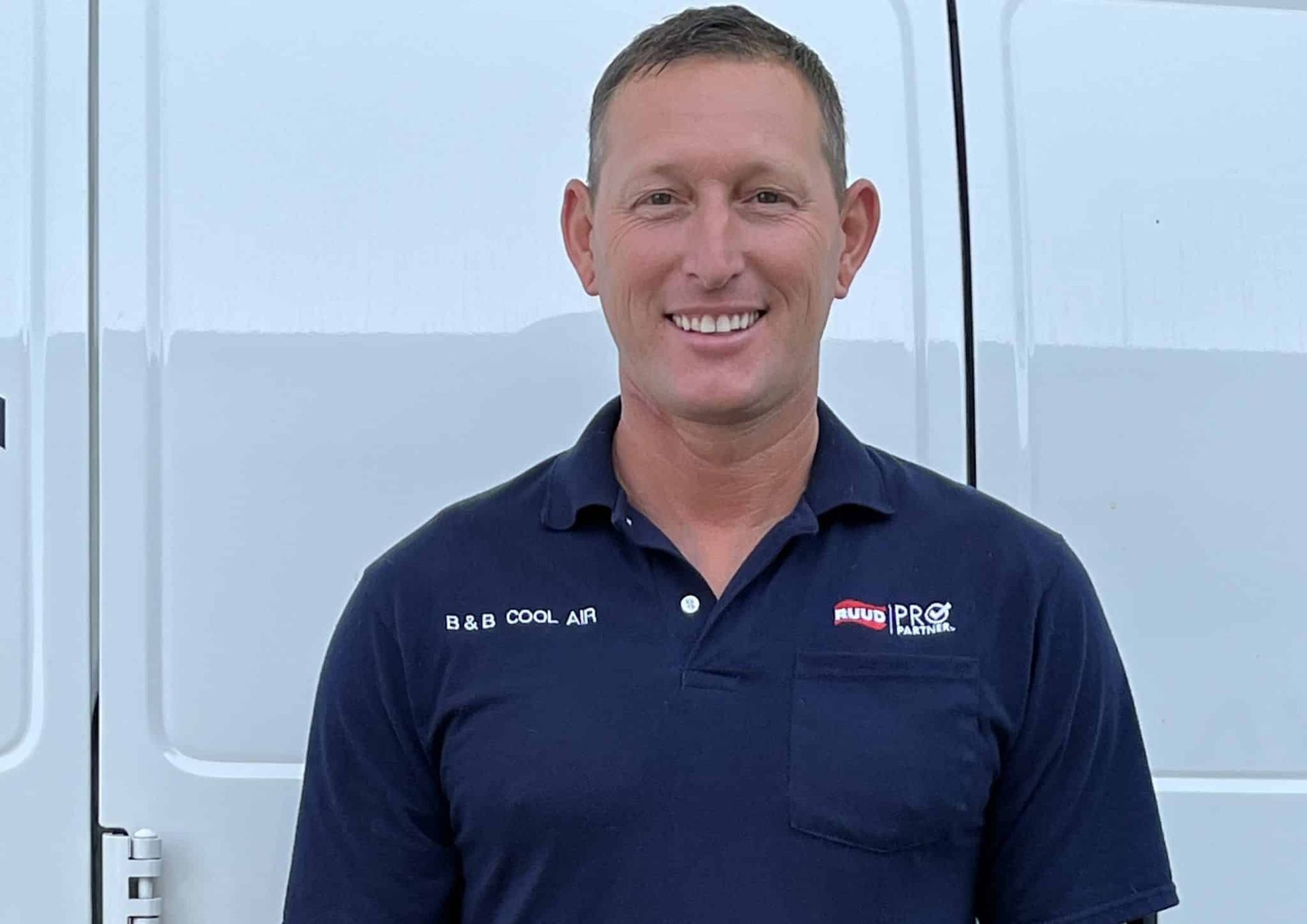 Man in blue polo shirt smiling in front of a white vehicle.