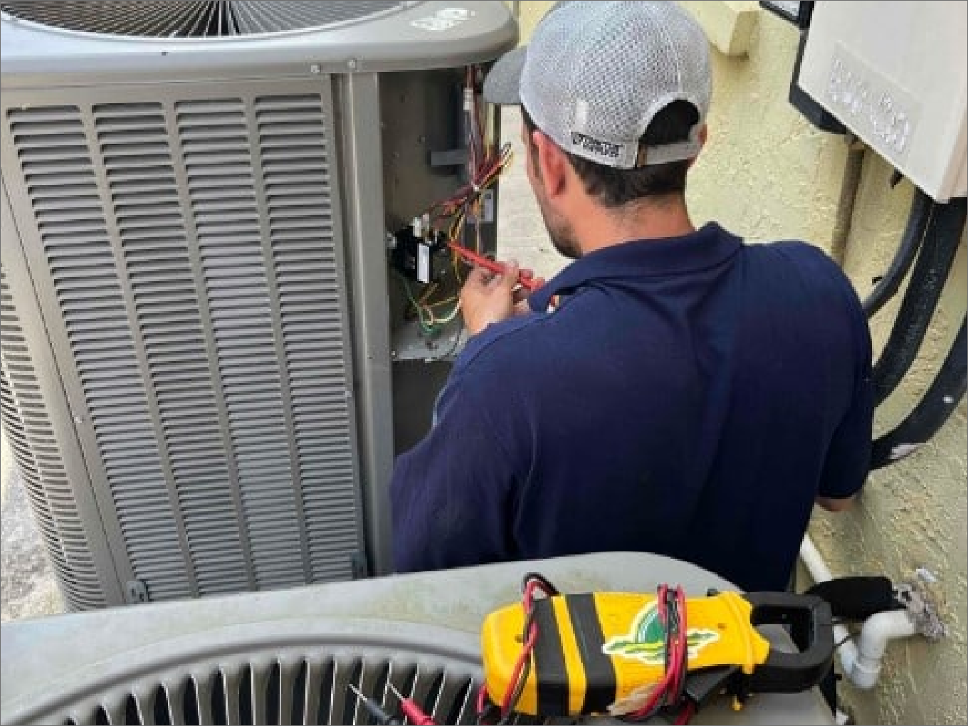 HVAC technician in a blue shirt and cap working on an AC unit outside.