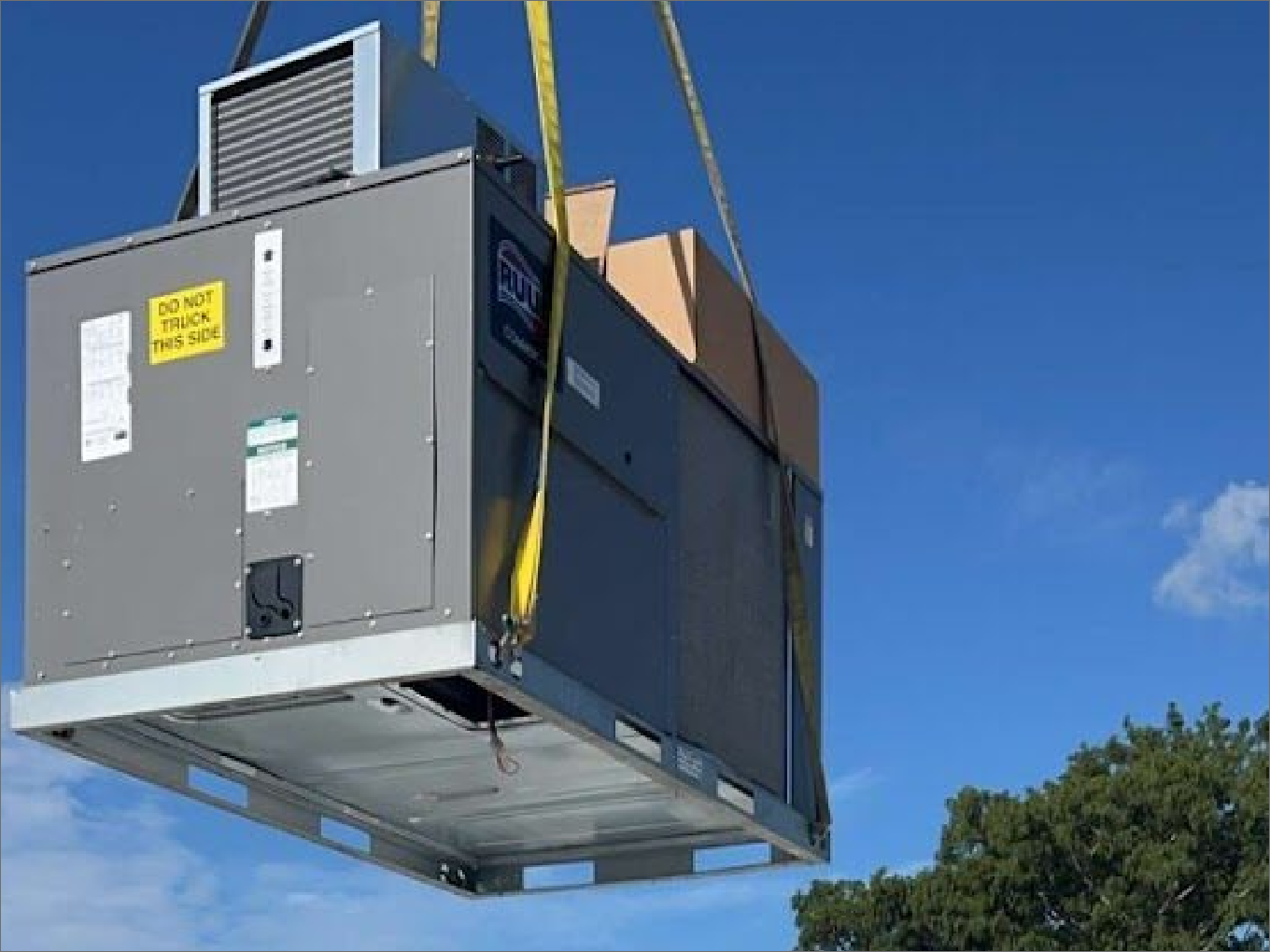 HVAC unit lifted by crane with yellow straps against a blue sky, tree in the corner.