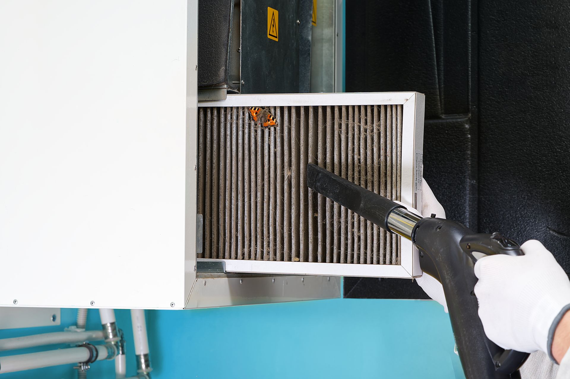 A gloved hand cleans a dirty air filter with a vacuum nozzle near an air conditioning unit.