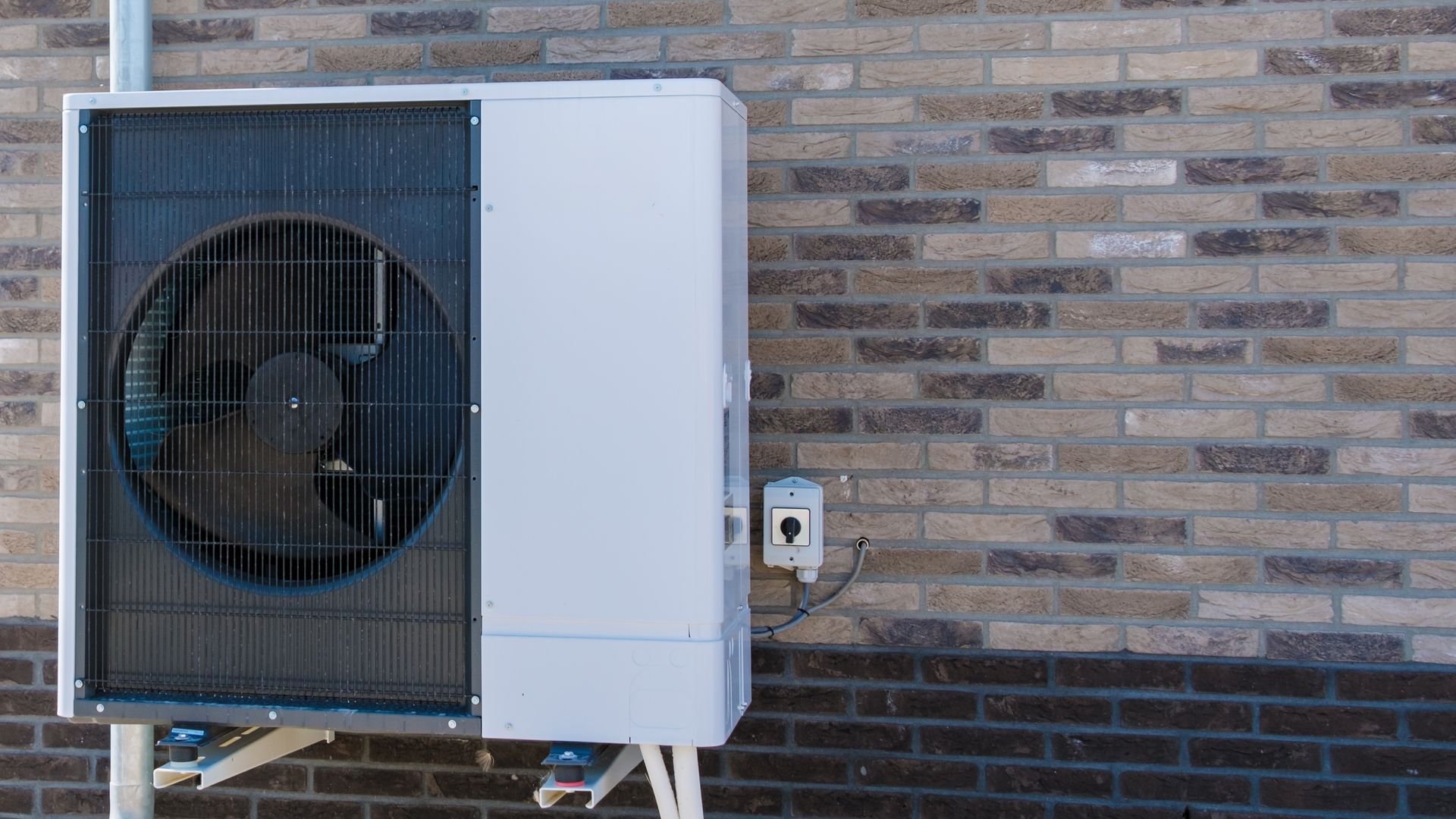 Outdoor heat pump unit mounted on a brick wall, with a fan visible behind a protective grille.