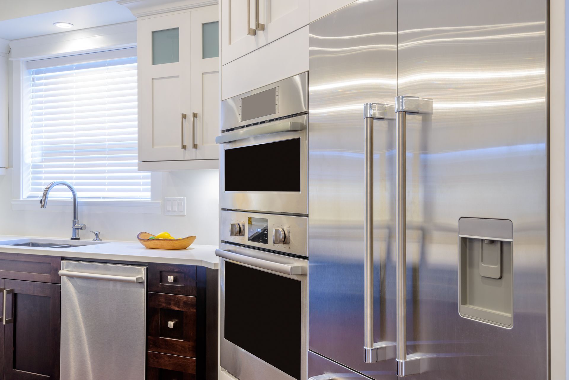Modern kitchen with stainless steel refrigerator, ovens, white cabinets, and a sink by a window.
