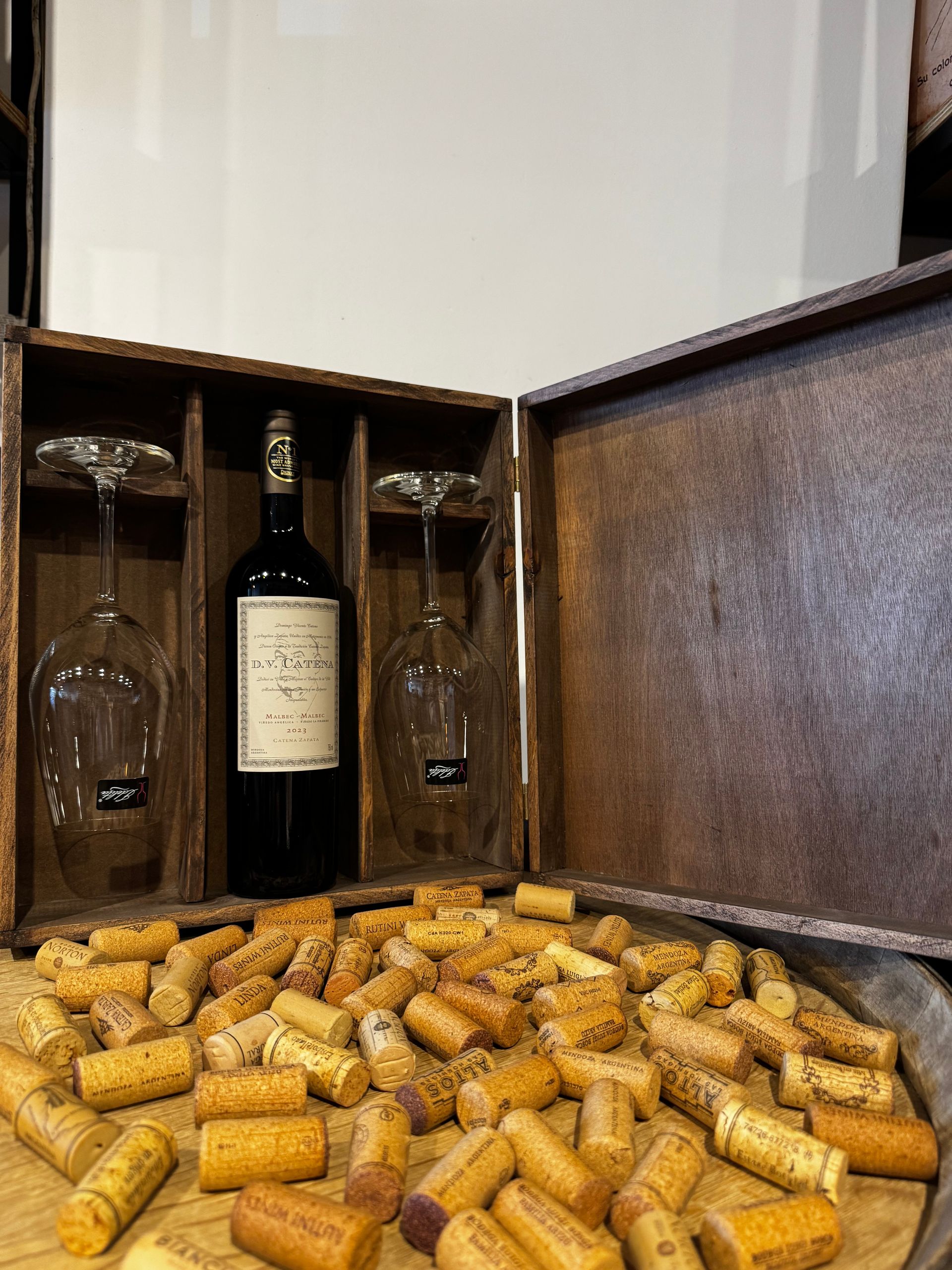 Botella de vino y copas en una caja de madera, con corchos derramándose sobre una bandeja.