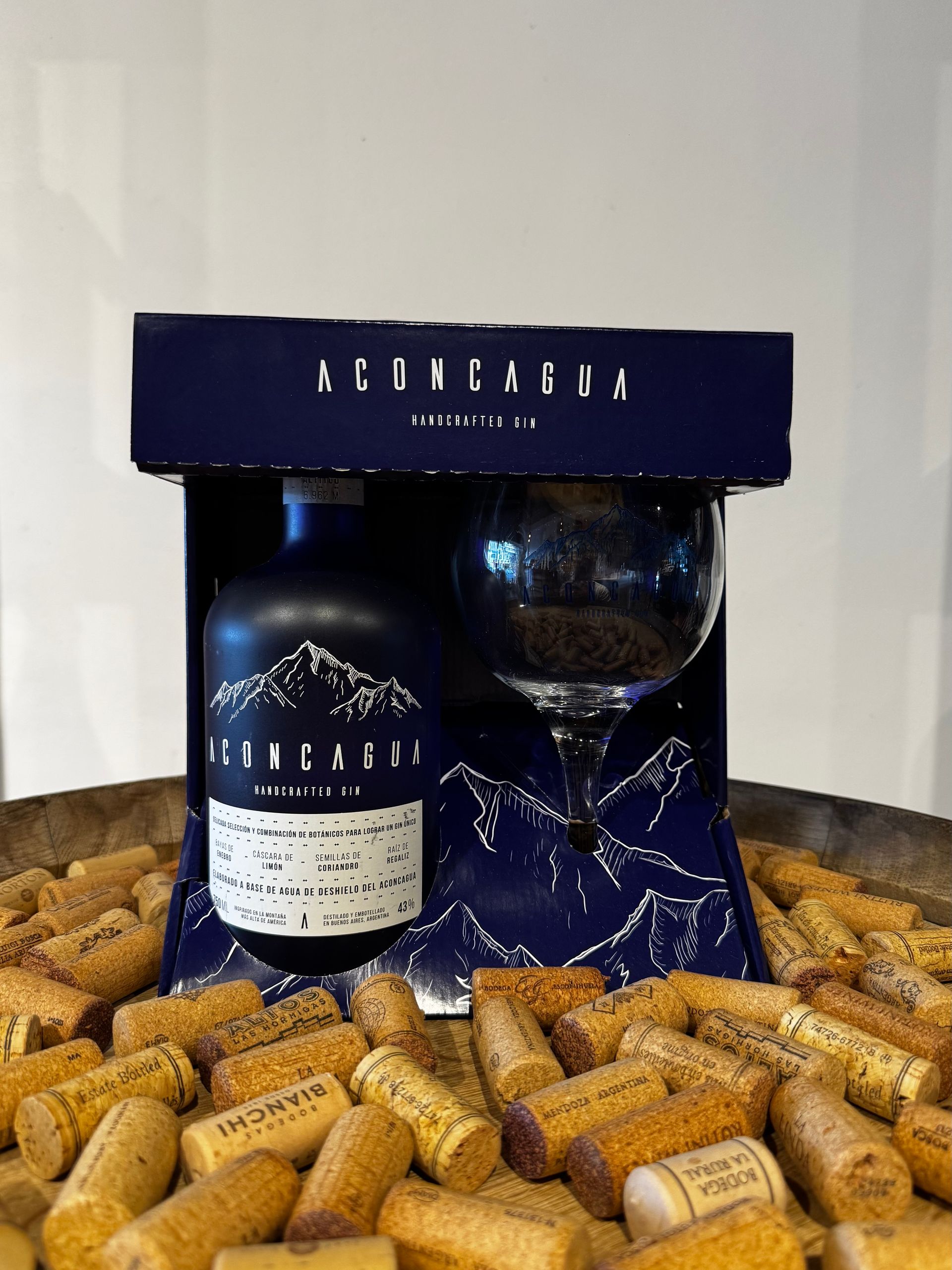 Set de regalo en caja azul, botella negra y copa de vino con su contenido, sobre lecho de corchos.