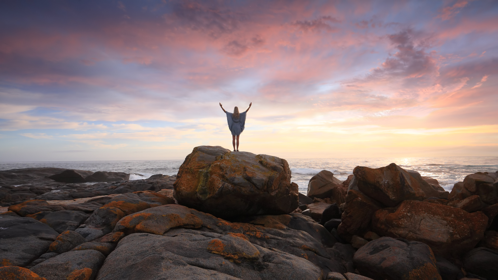 Una persona está parada en la cima de una roca cerca del océano con los brazos en el aire.