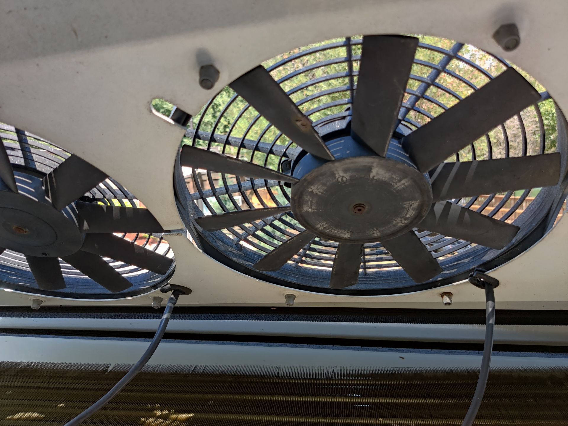 Two large black fan blades behind a metal grille, mounted on a white surface. Wires and outdoor scenery are visible.
