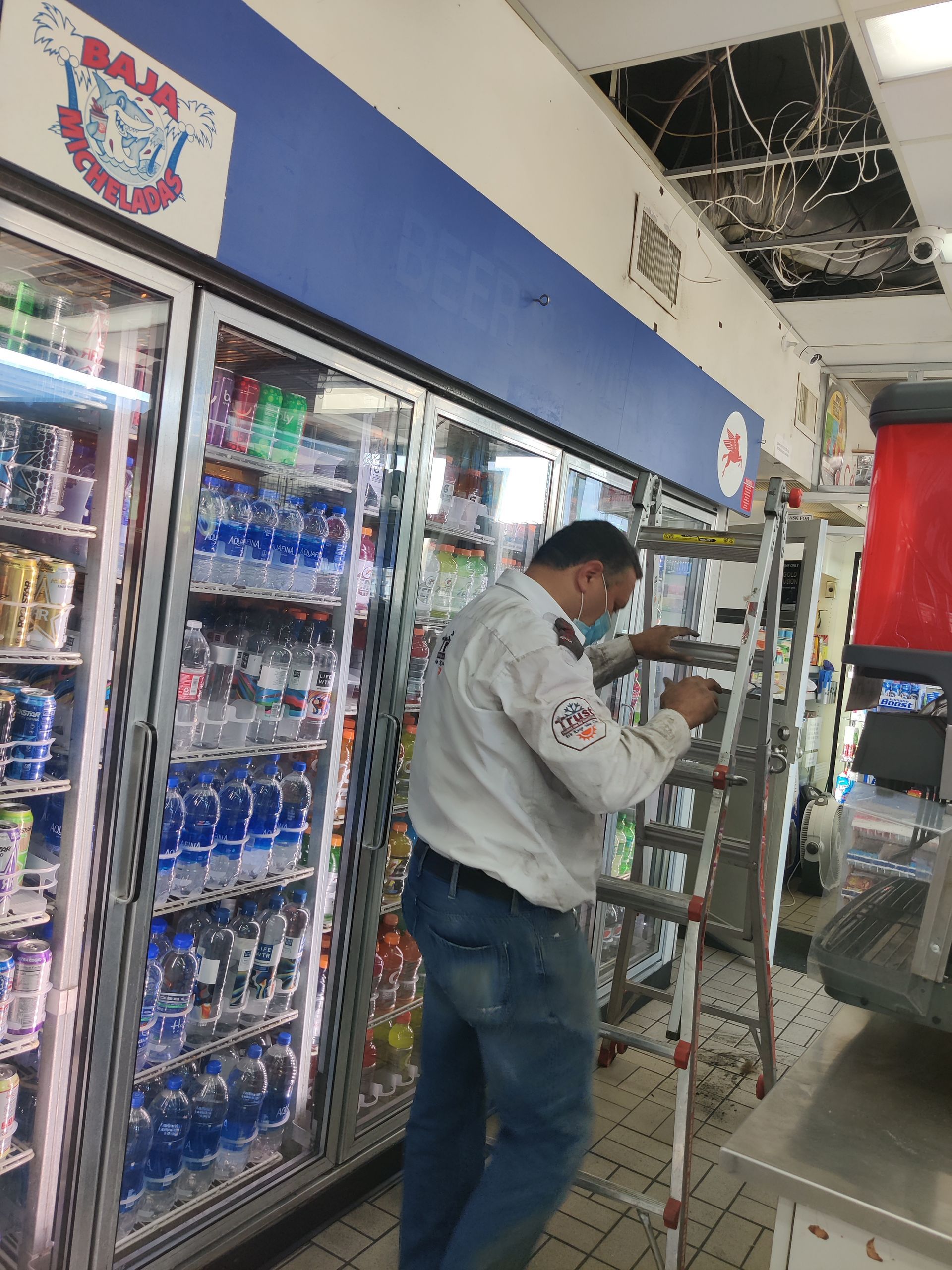 Person on a ladder working above store coolers; ceiling damage visible.
