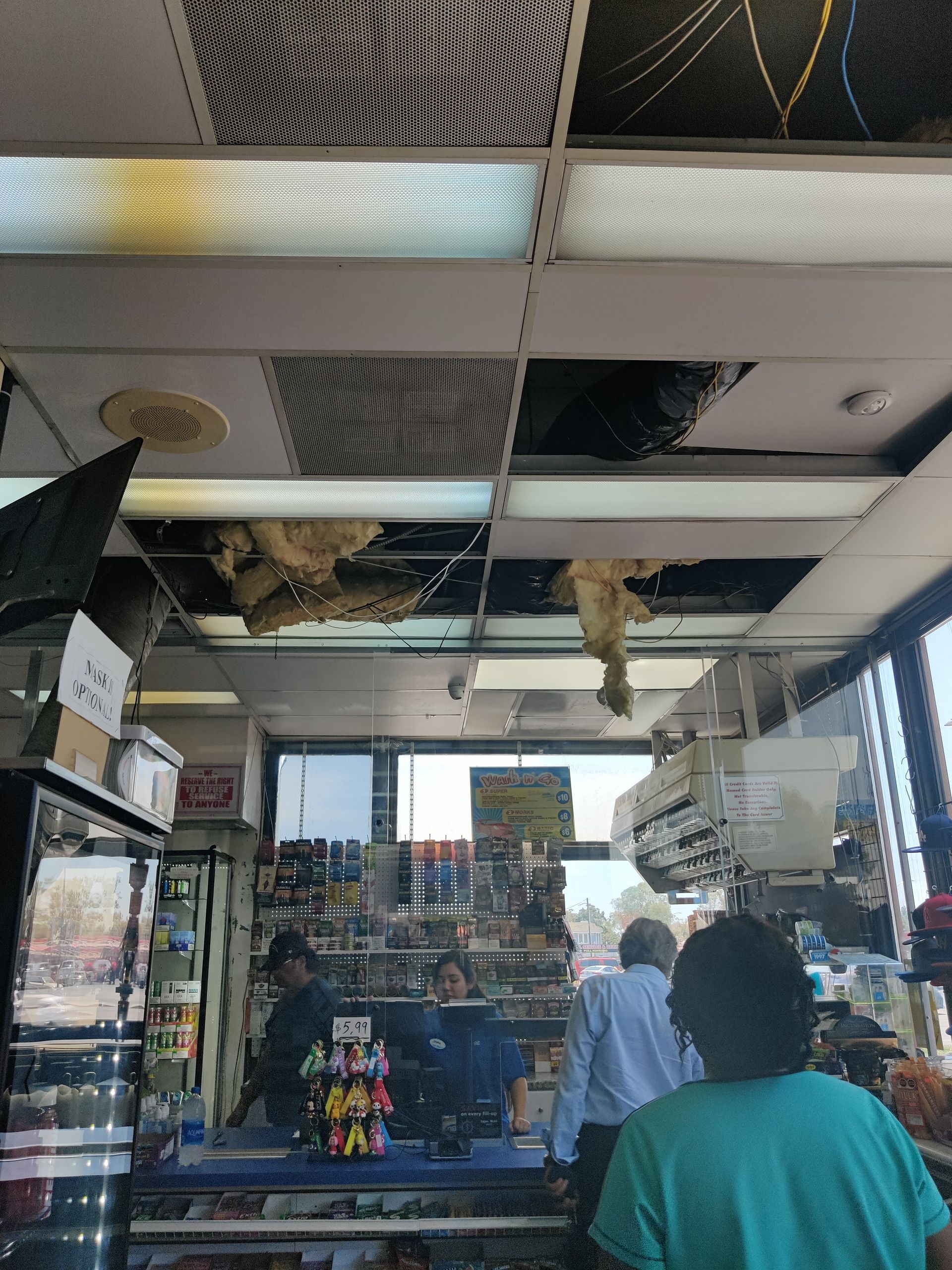 Interior of a convenience store with damaged ceiling tiles. Customers at the counter, shelves of goods visible.