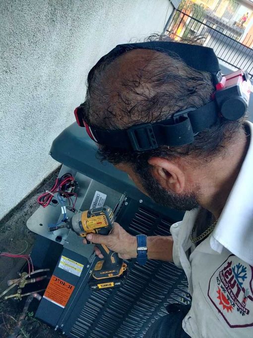 HVAC technician using a drill on an air conditioning unit. Outdoors, wearing safety glasses and a uniform.