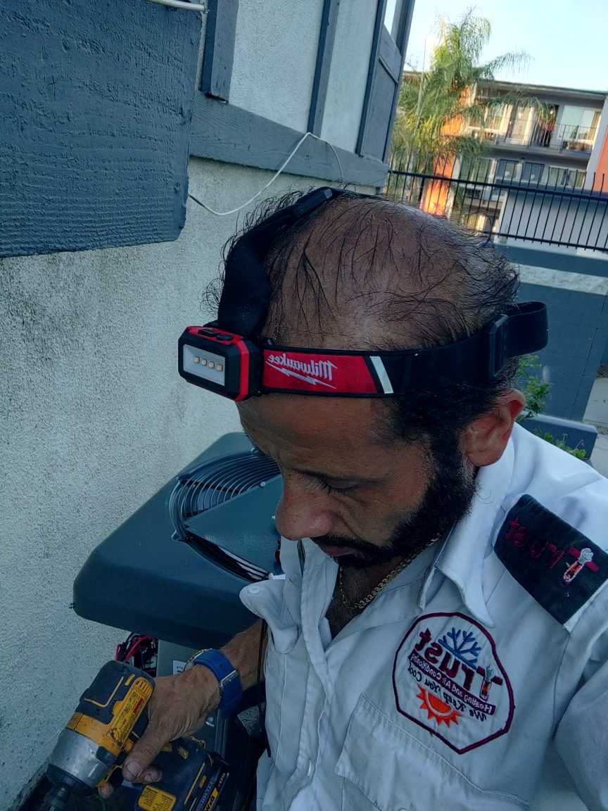 Man in a uniform wearing a headlamp, working on an air conditioner with a drill.