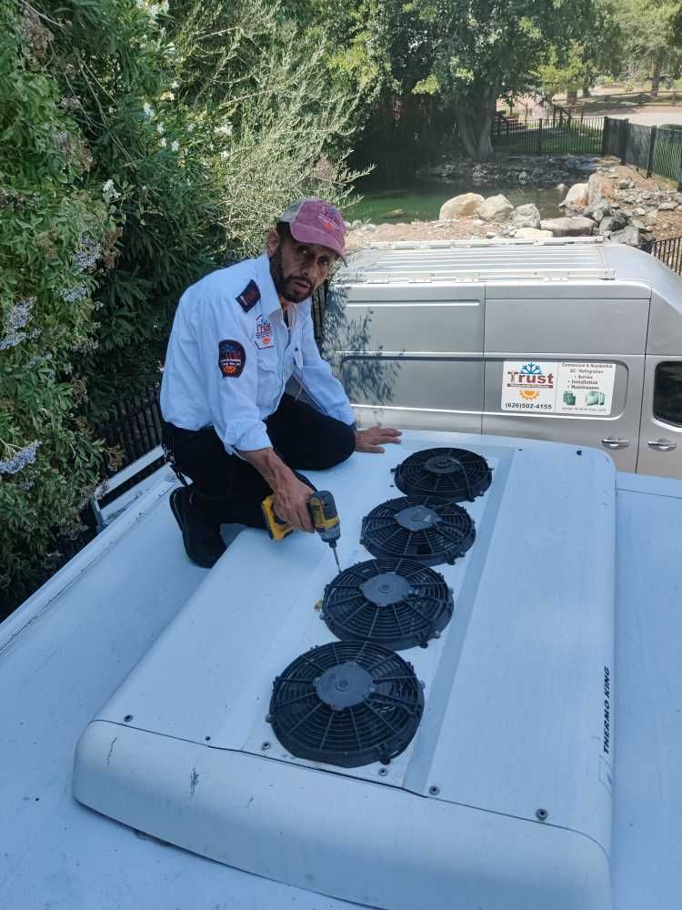 Man working on rooftop air conditioning unit, using a drill. Outdoor setting, bright daylight.