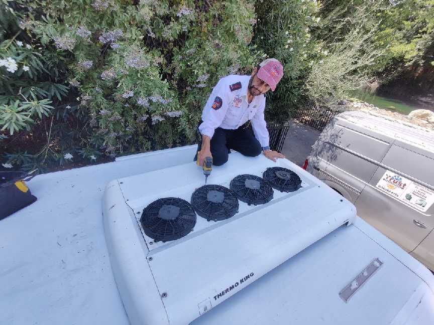 A person in work attire kneels on a white vehicle roof, using a drill near black fan vents.