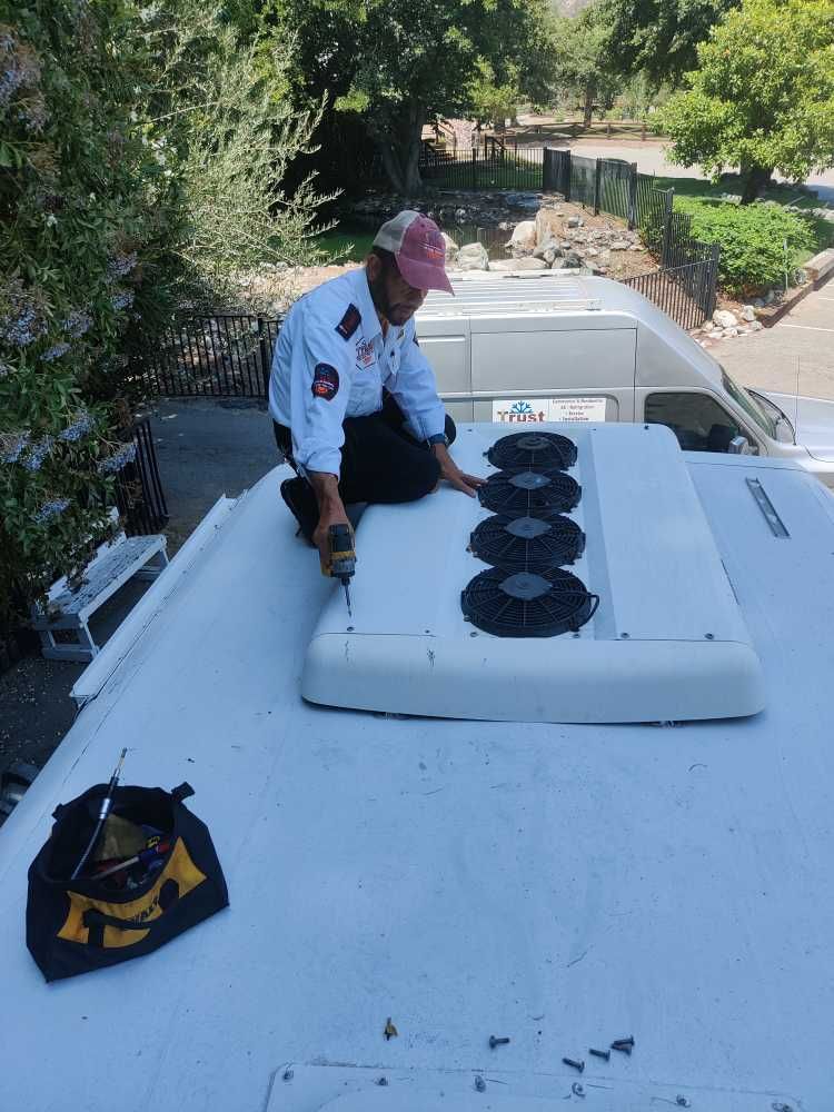 Man on a vehicle roof repairing an air conditioner unit, holding a drill.