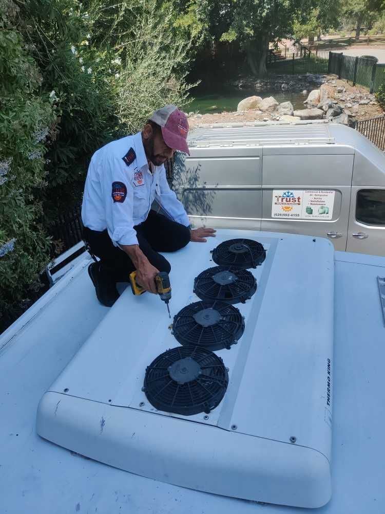 A person in work attire uses a drill on an air conditioning unit atop a vehicle.