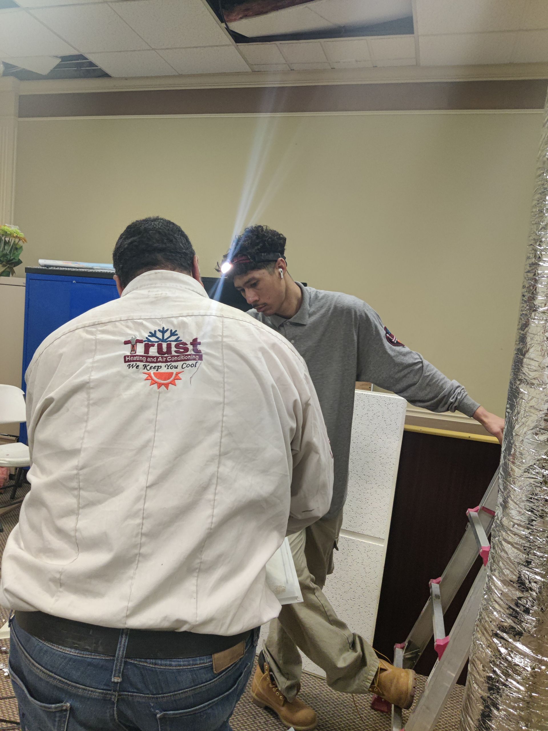 Two people repairing a ceiling. One on a ladder, the other standing near it. Beige walls and damaged ceiling visible.