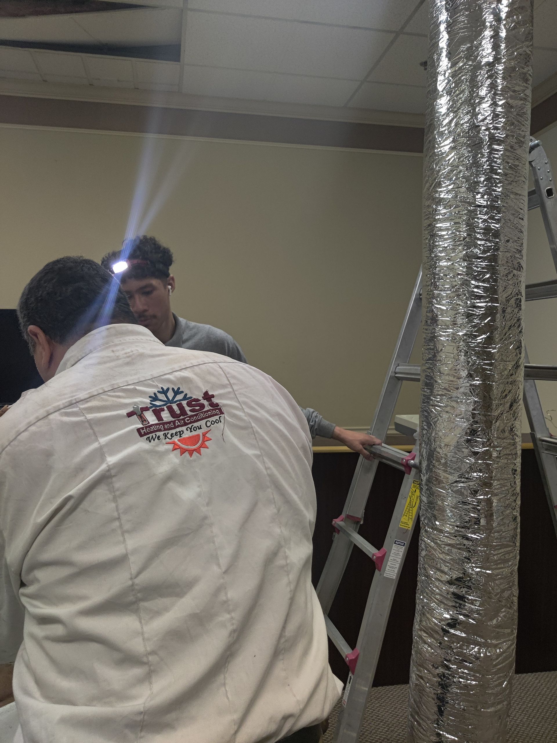 Two people inspecting ductwork in a room. One on a ladder, using a headlamp.