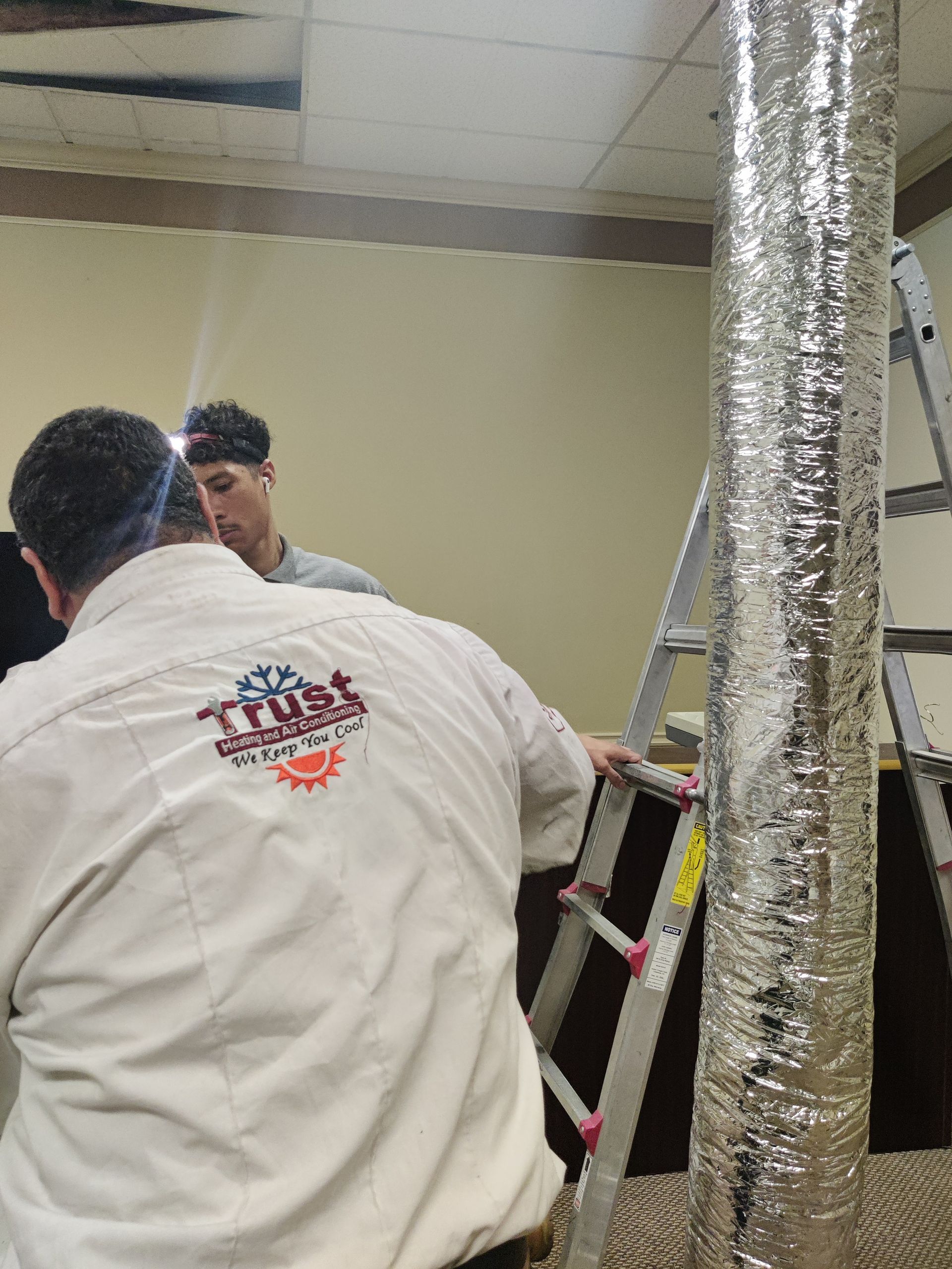Two people working on a duct, one on a ladder, in an office with beige walls. The duct is wrapped in silver foil.