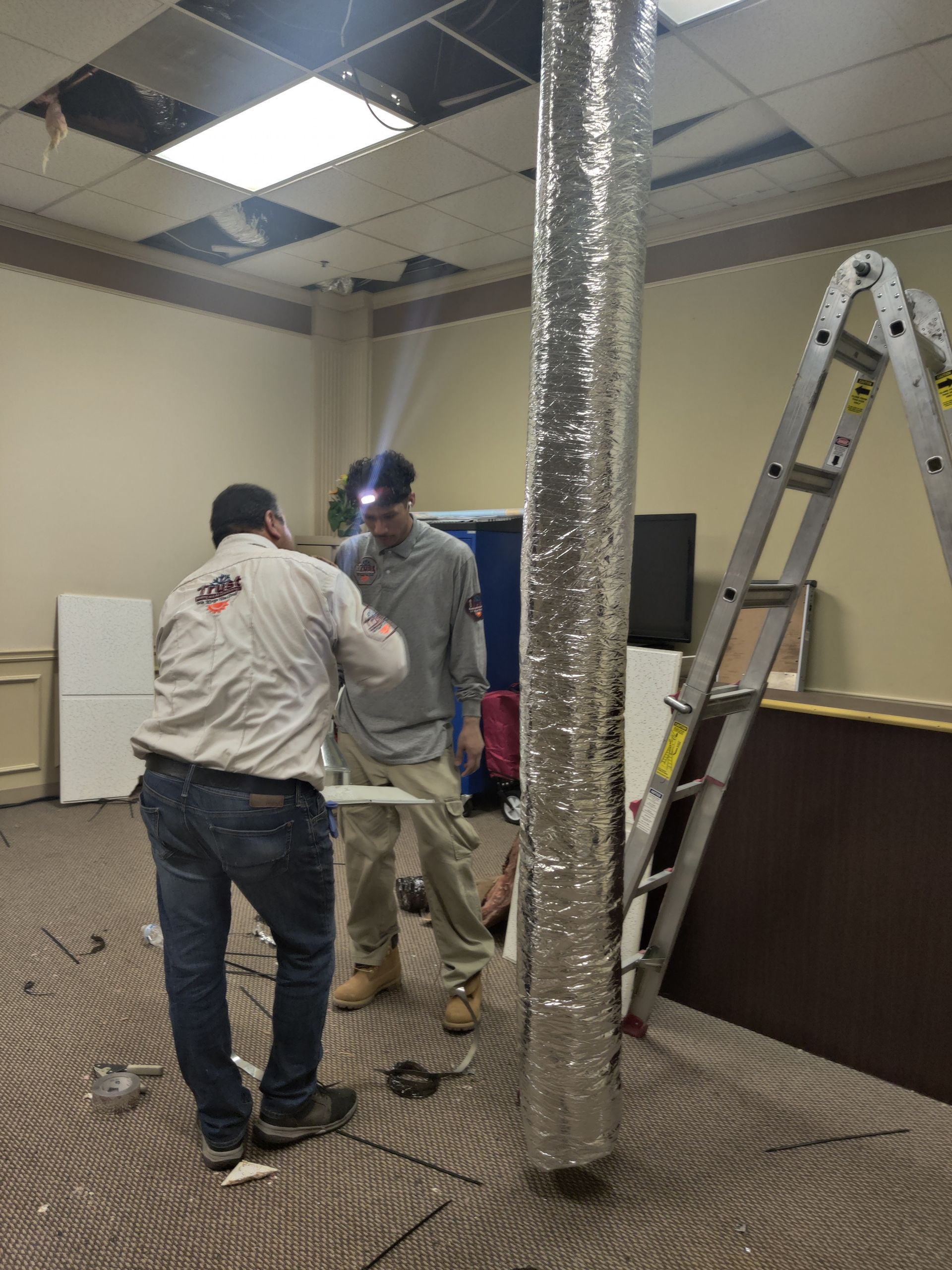 Two men working on ceiling, inspecting a silver duct. A ladder leans against the wall in a room.