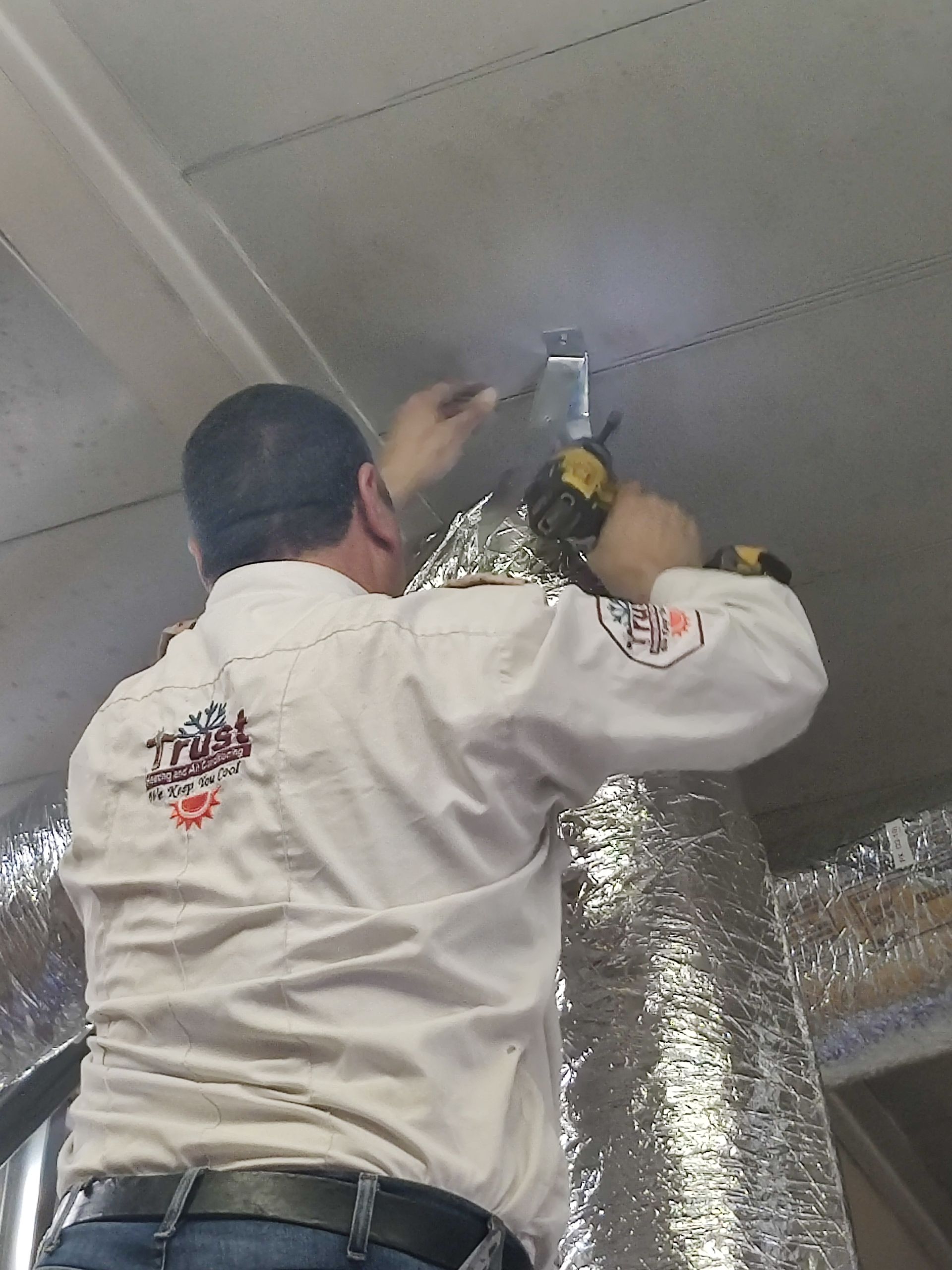 Person in a white shirt installing something on a ceiling with a drill; reflective insulation is visible.