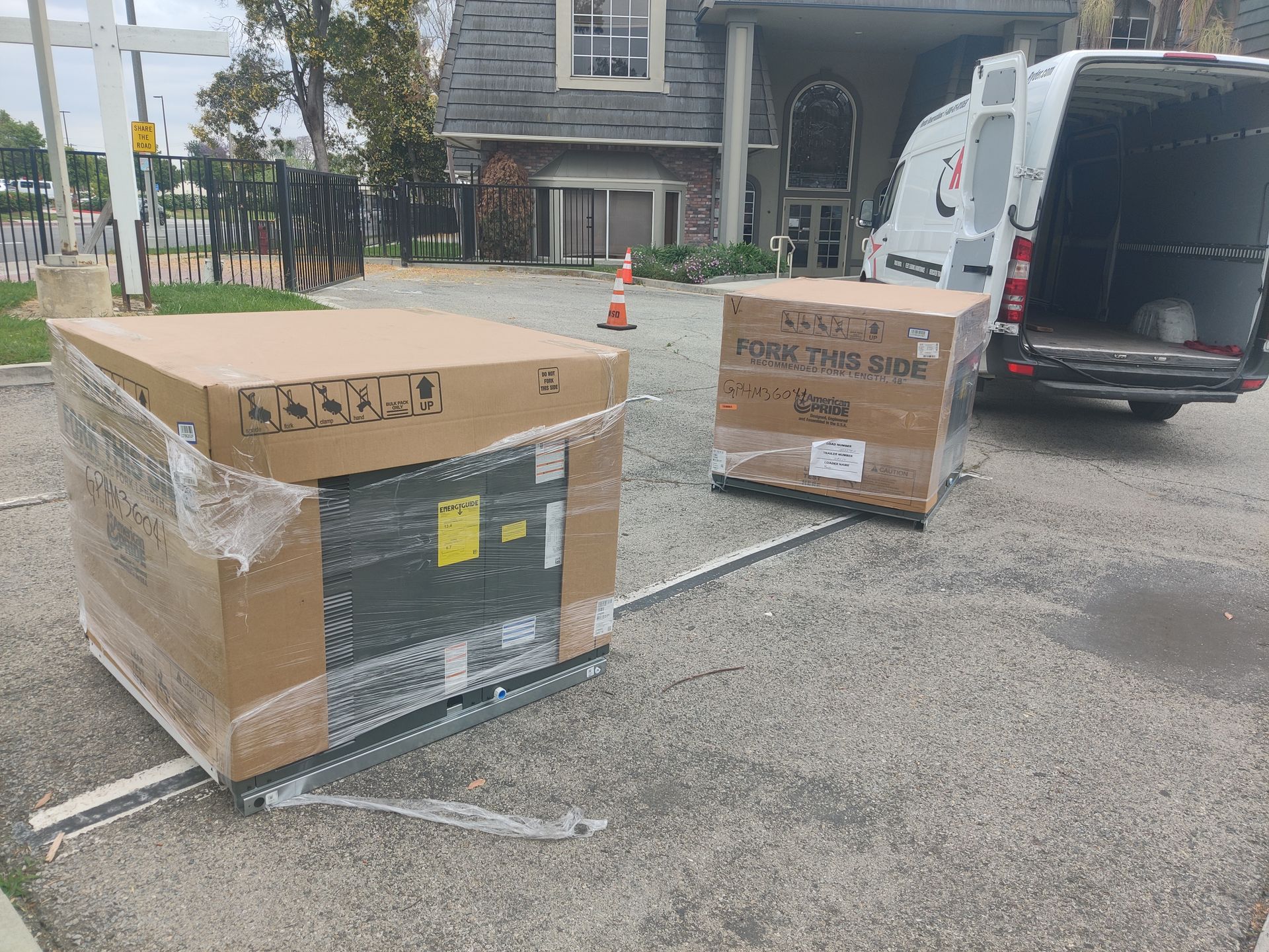 Two large, boxed items on pallets in a parking area, near a delivery van with an open door.