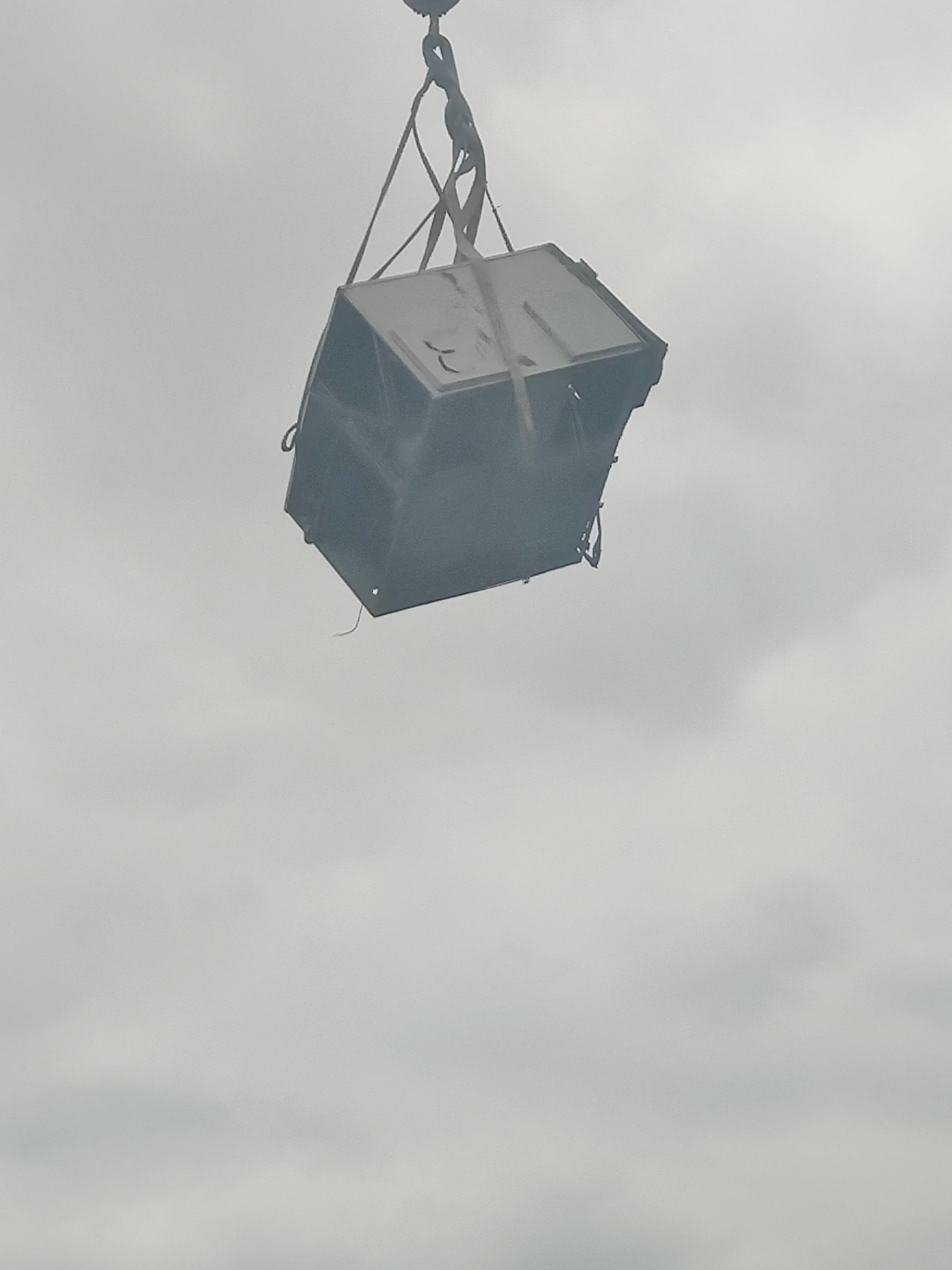 A box-like object suspended in the air by a crane against a cloudy sky.