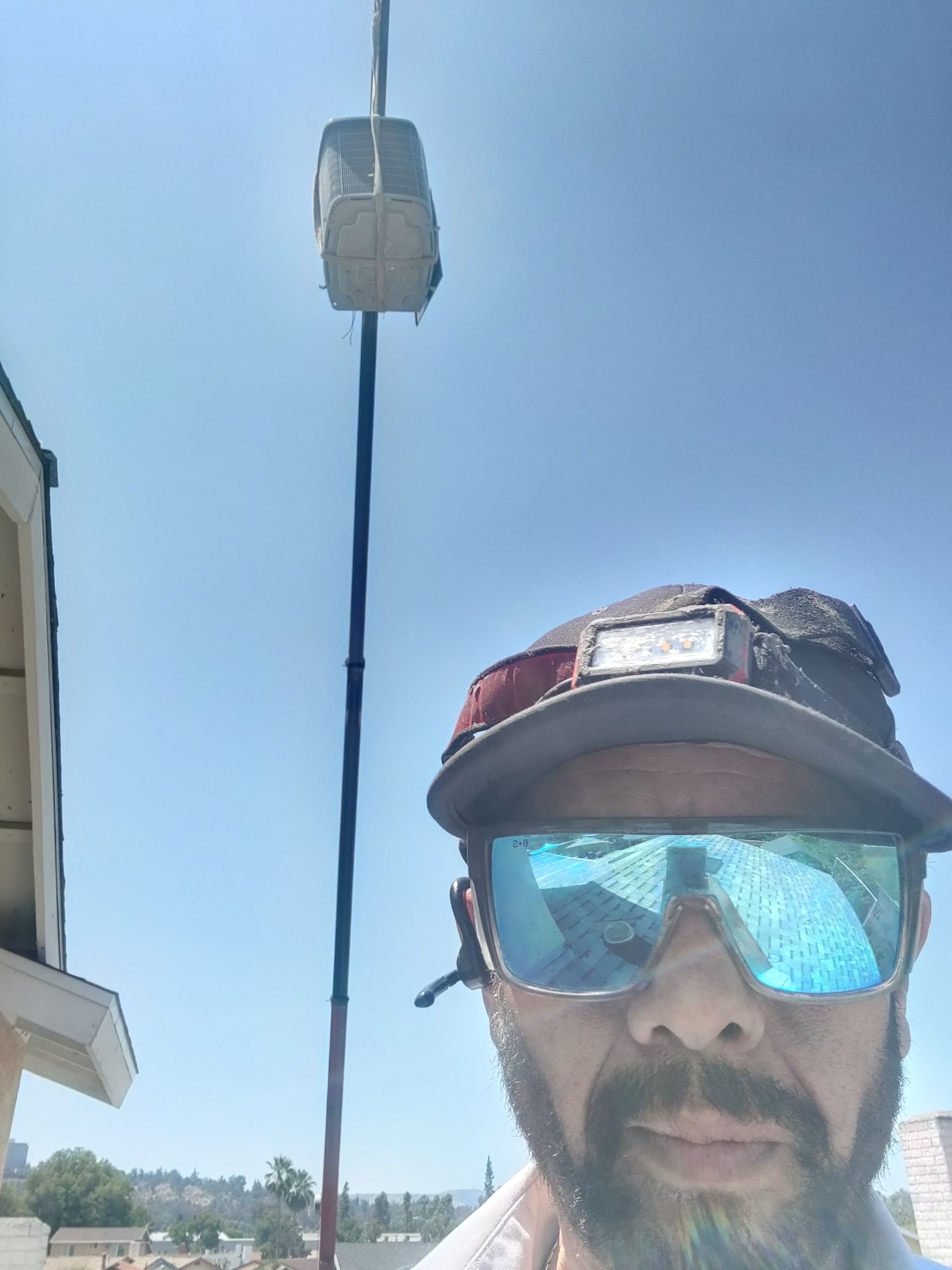 Man in sunglasses on roof, looking at camera. Tall pole with a box attached to it. Blue sky.