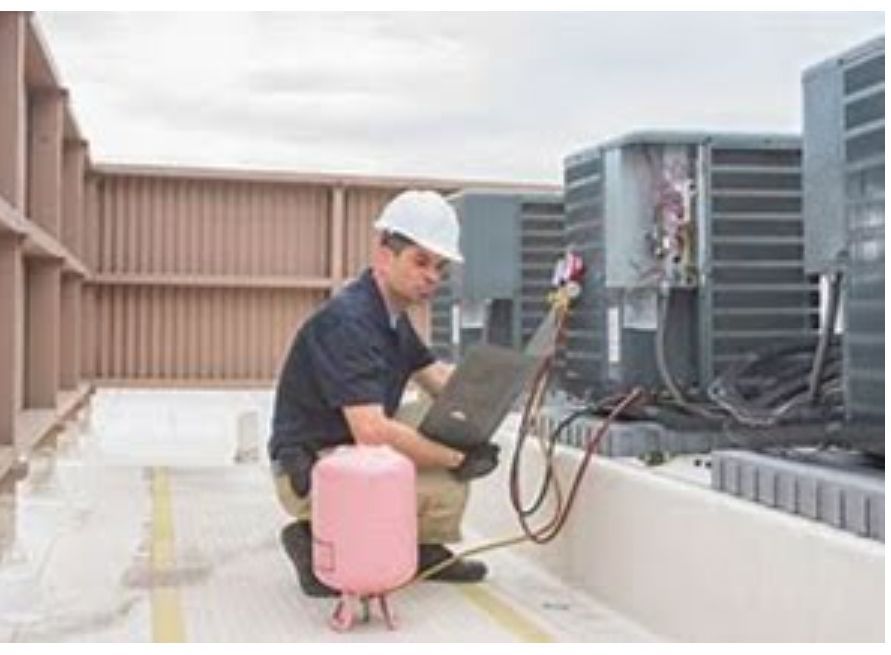 HVAC technician kneels on a rooftop, using a laptop and gauges to service an air conditioning unit.