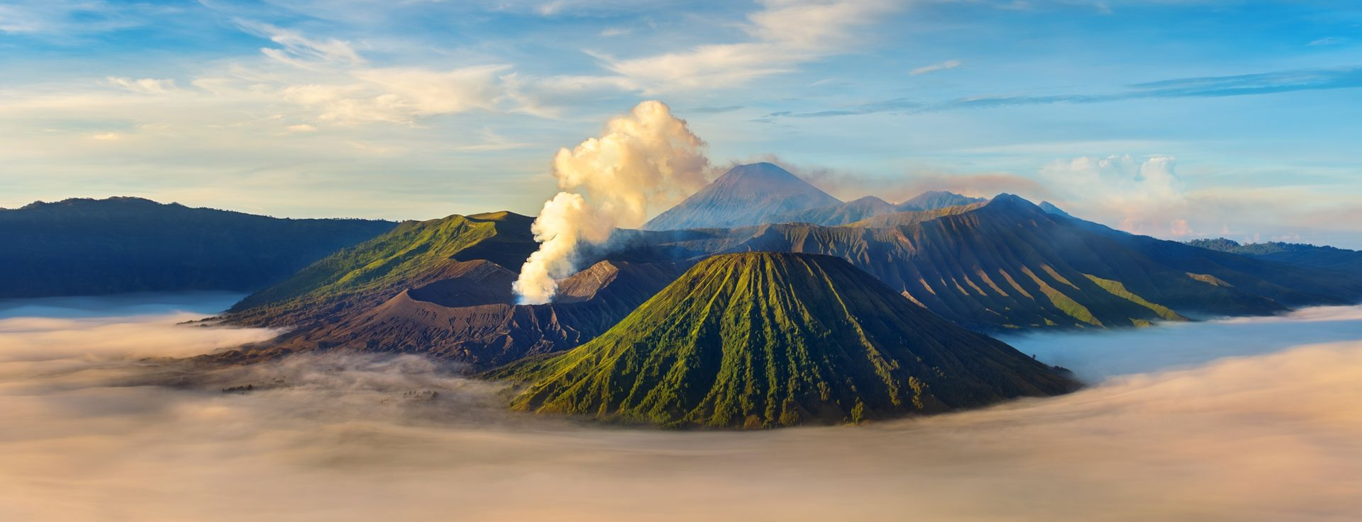 Volcanic landscape with green cones and a plume of smoke, shrouded in fog under a blue sky.