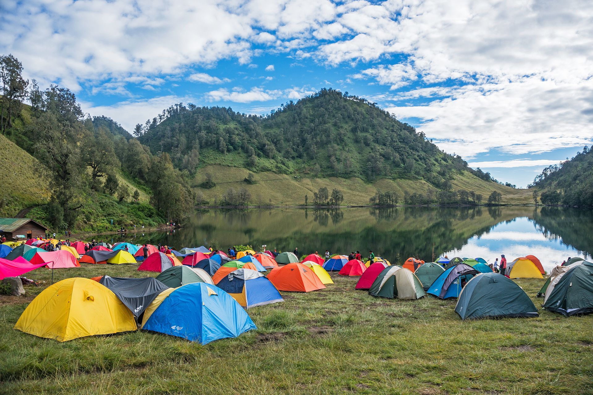 Camping tents set up on a grassy field by a lake, mountains in the background, blue sky with clouds.