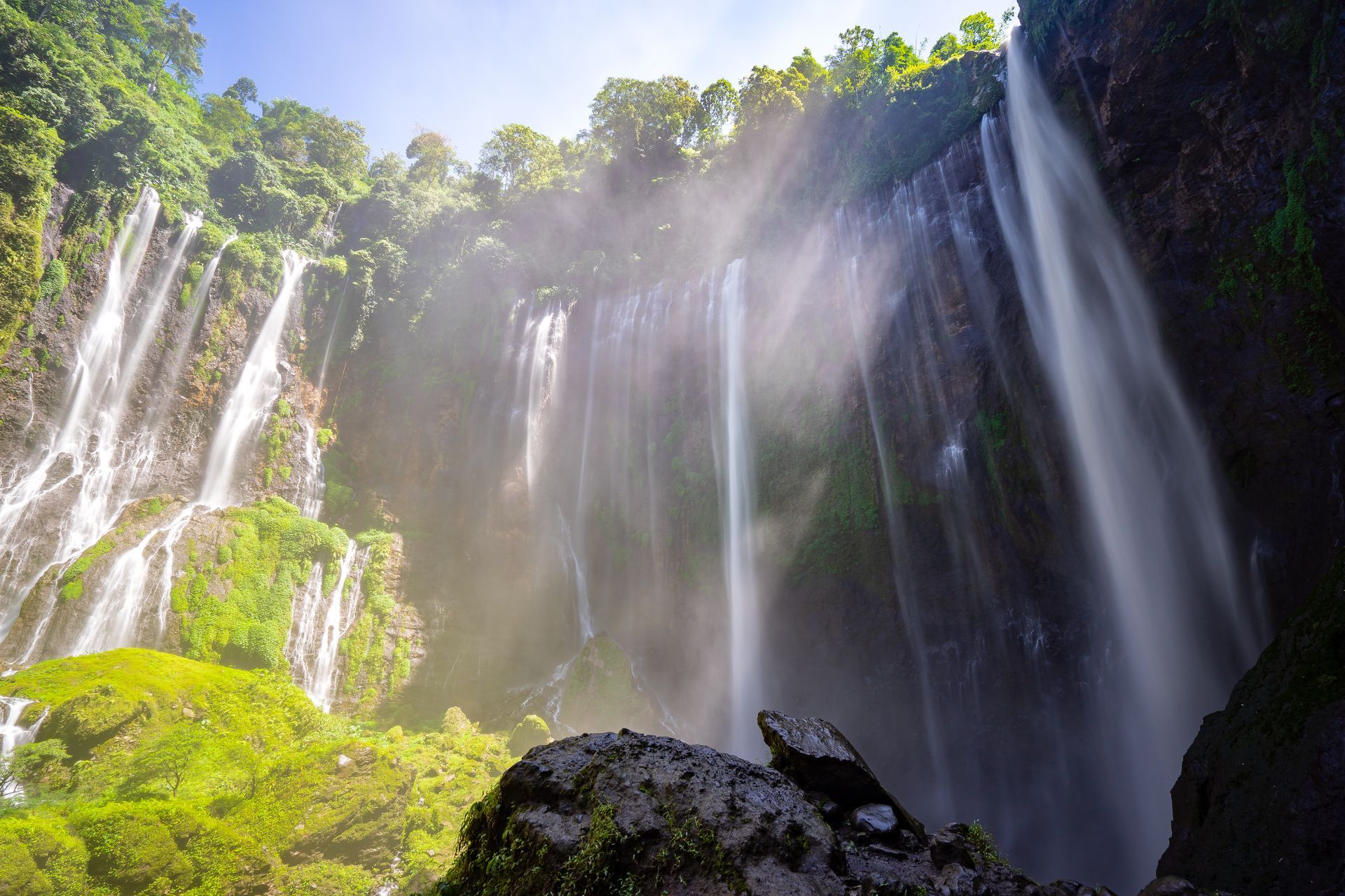 Waterfall cascading down a cliff, surrounded by lush green vegetation. Sunlight creates a misty haze.