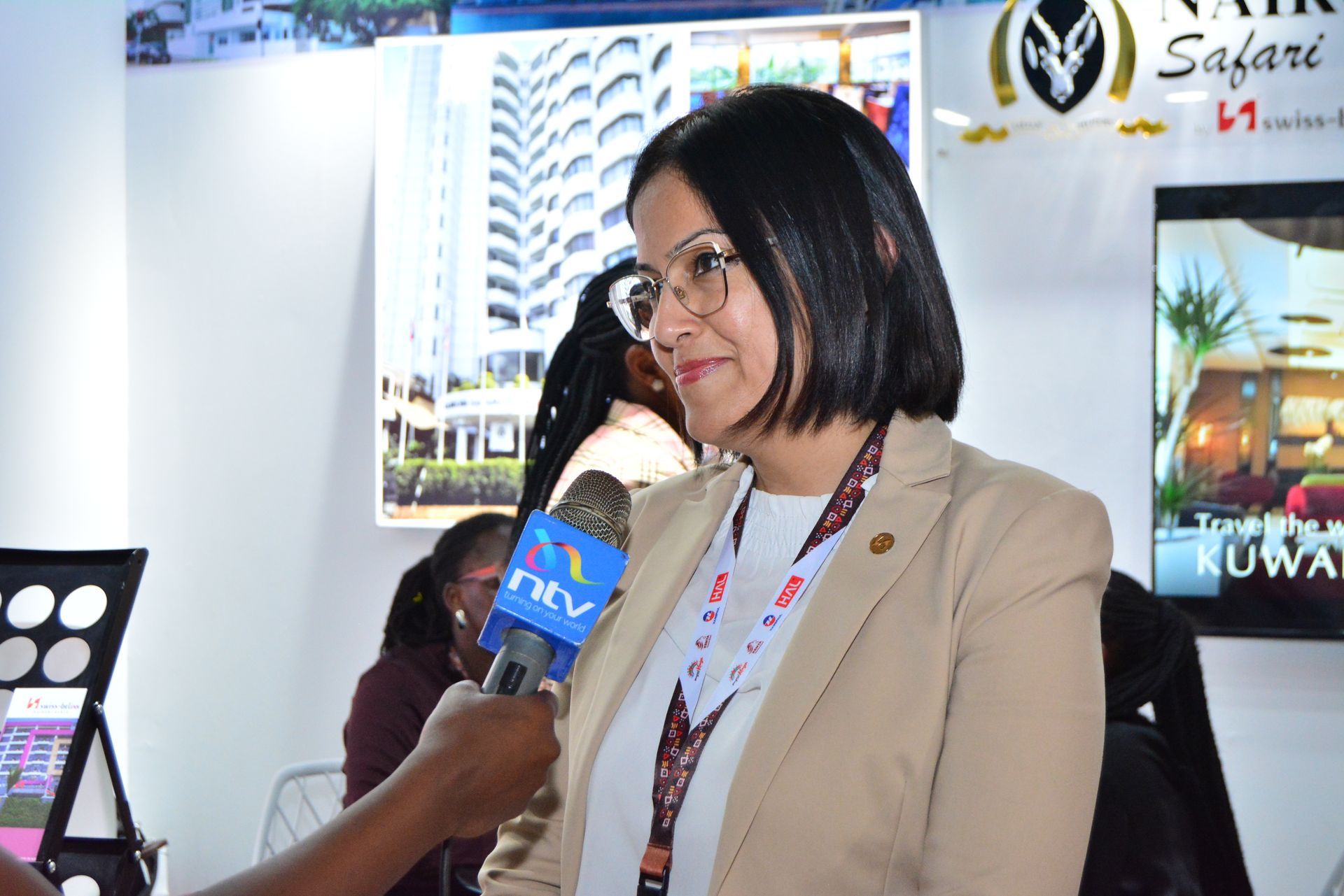 Woman being interviewed at a trade show; beige blazer, microphone, background with tourism logos.