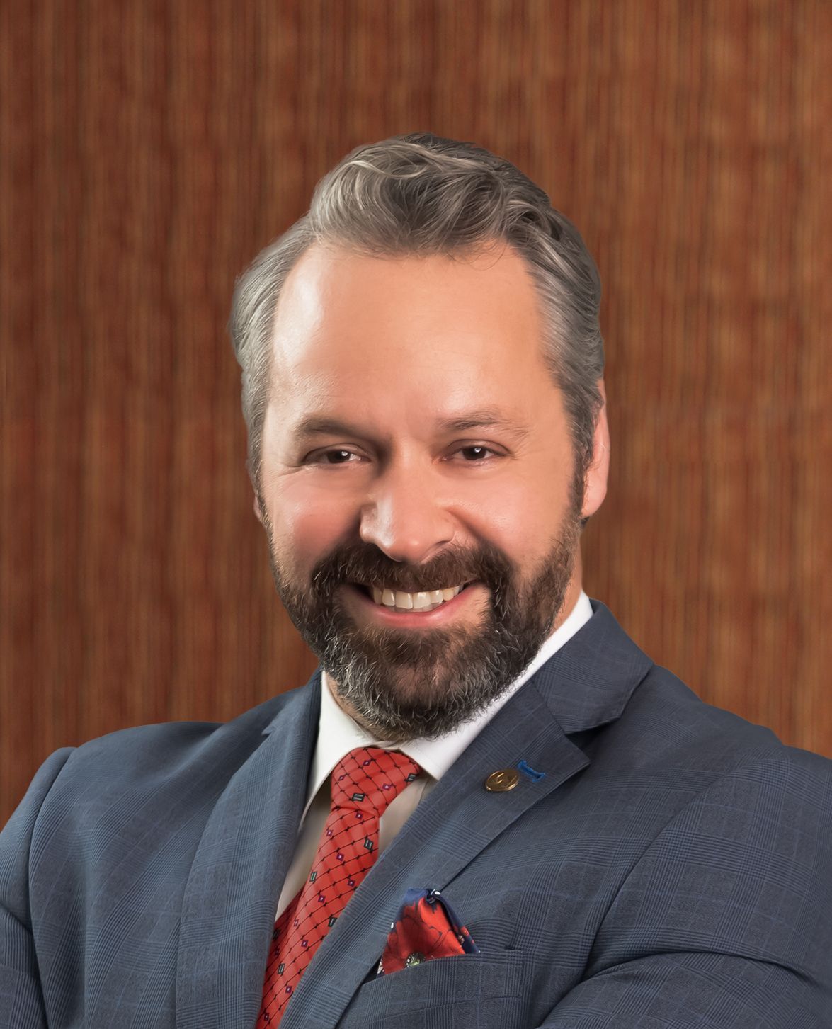 Man in a blue suit with a red tie smiles, set against a wooden background.