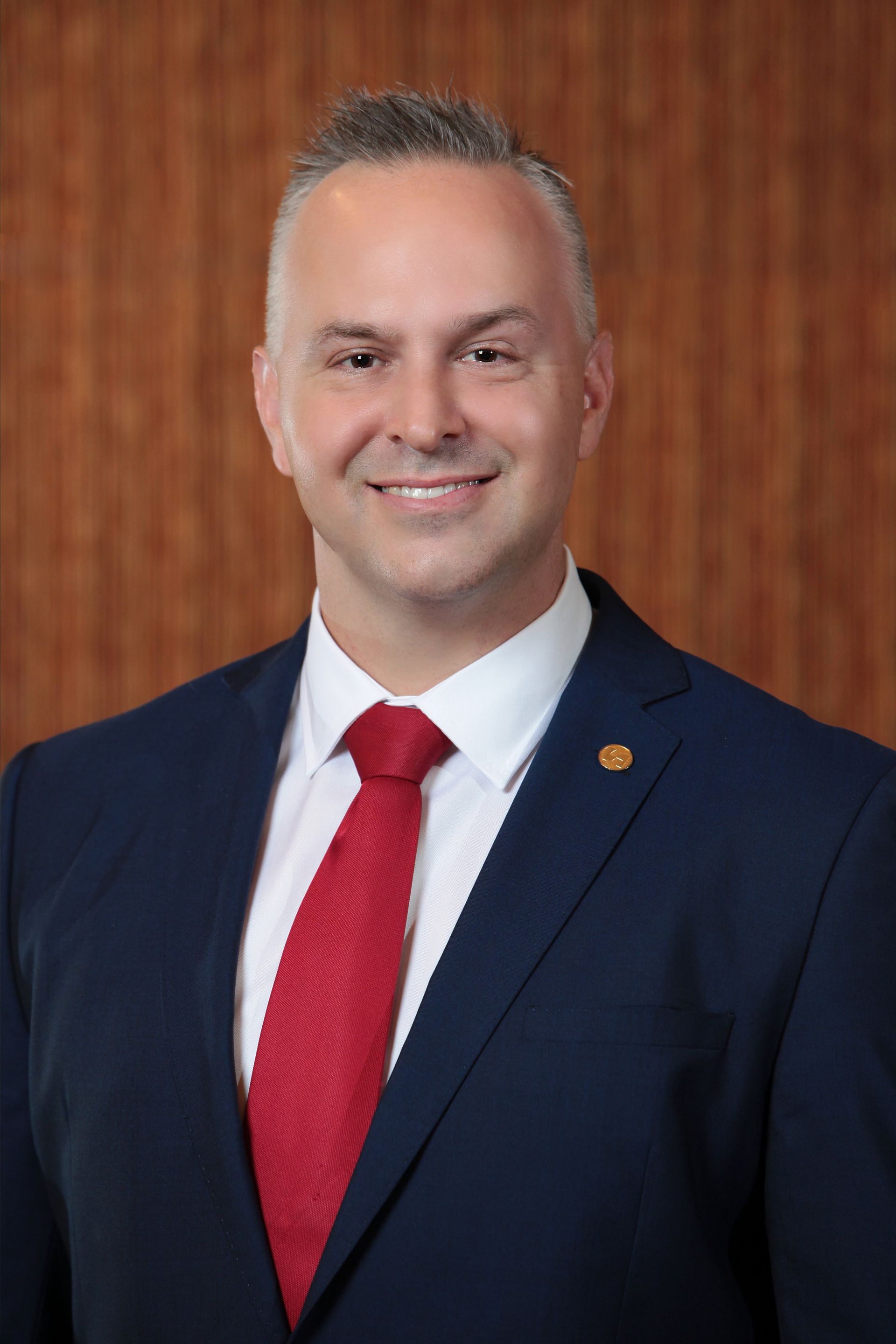 Man in a navy suit and red tie, smiling in front of a wood-paneled background.