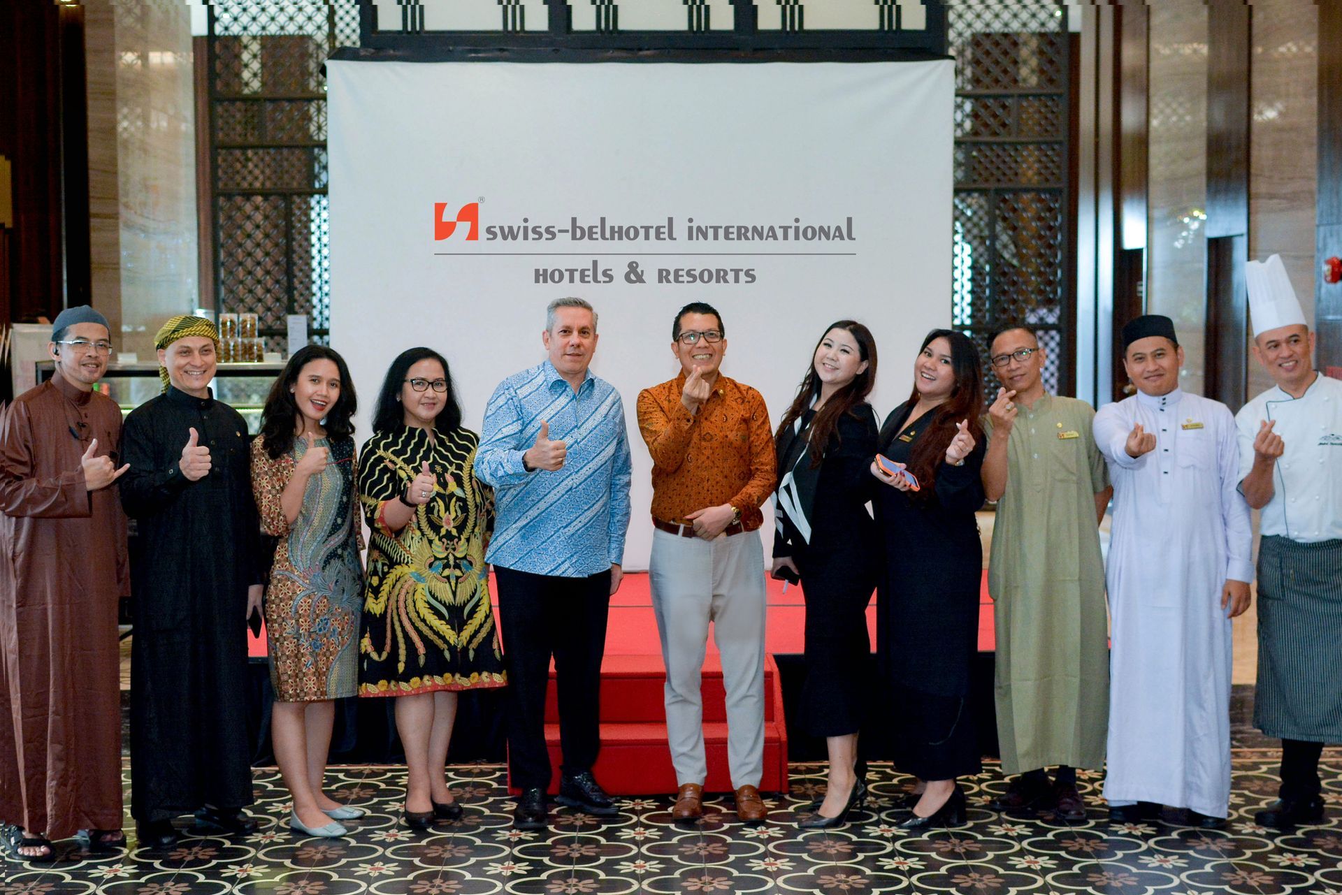 Group of people at Swiss-belhotel International event, thumbs up pose. White backdrop, red logo, indoor setting.