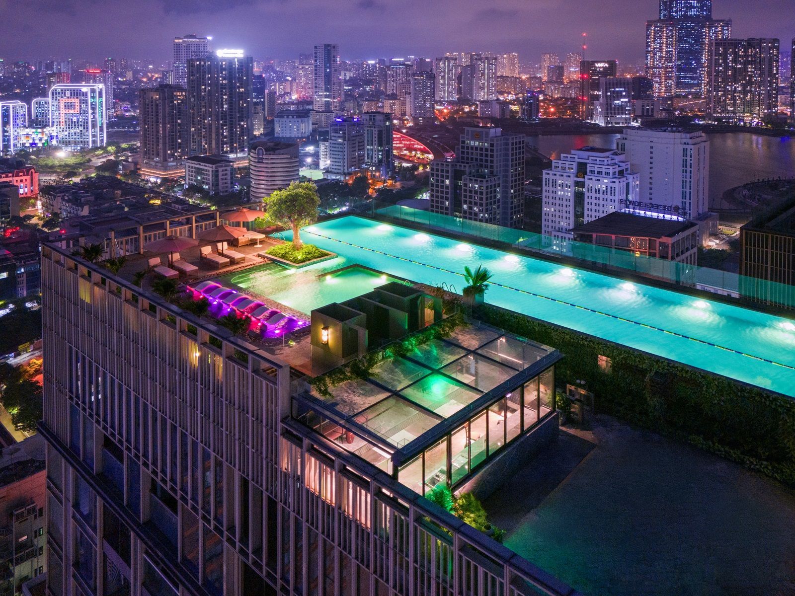 Rooftop pool at night with city skyline. Long pool glows turquoise, surrounded by modern buildings.