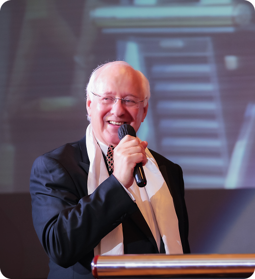 Man with microphone, smiling, wearing a black suit, white scarf, speaking at a podium.