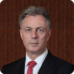 Man in suit and tie; serious expression, against a wood-paneled background.