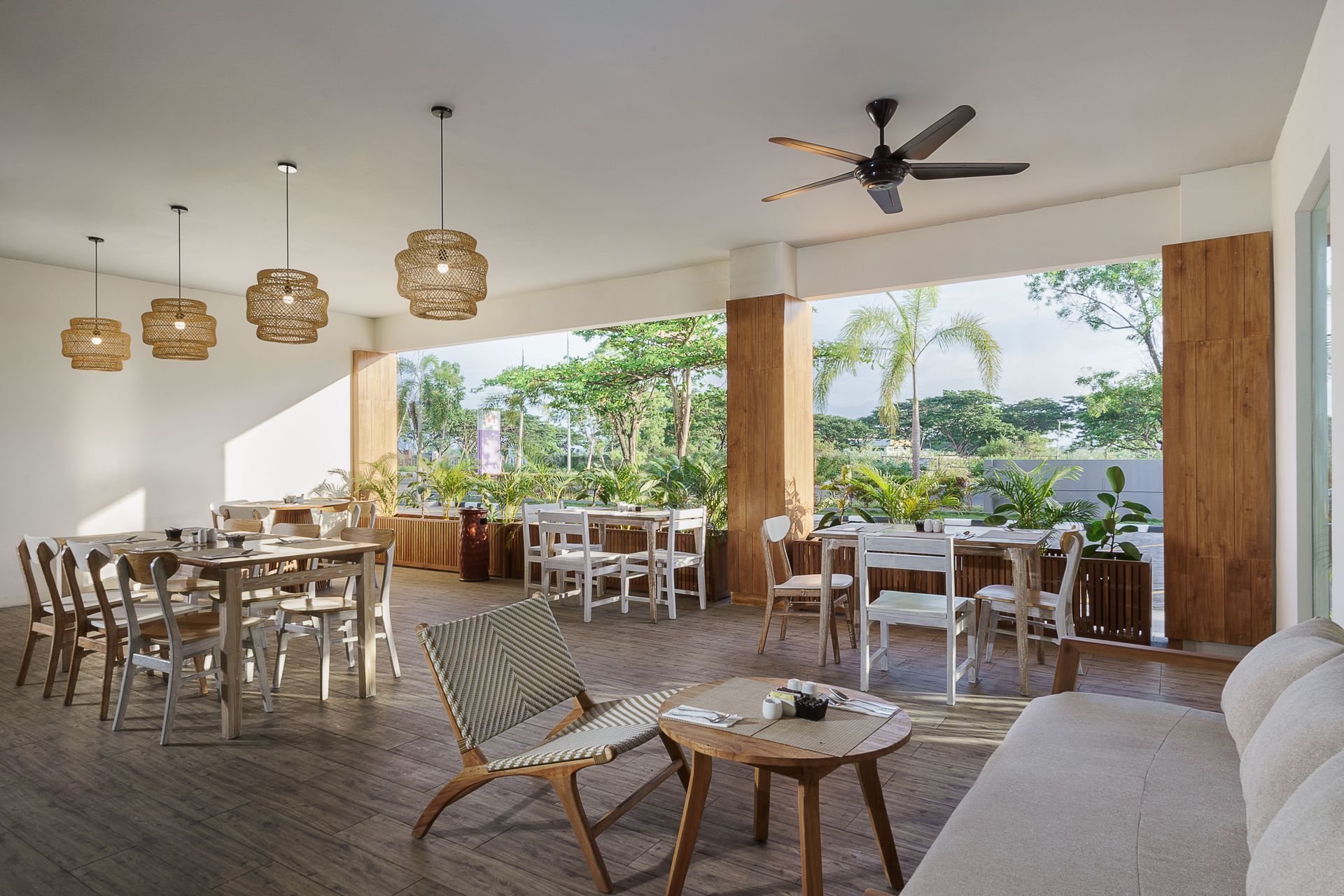 Outdoor dining area with tables, chairs, and a sofa. Lush greenery visible in the background. Wooden floors and pillars.
