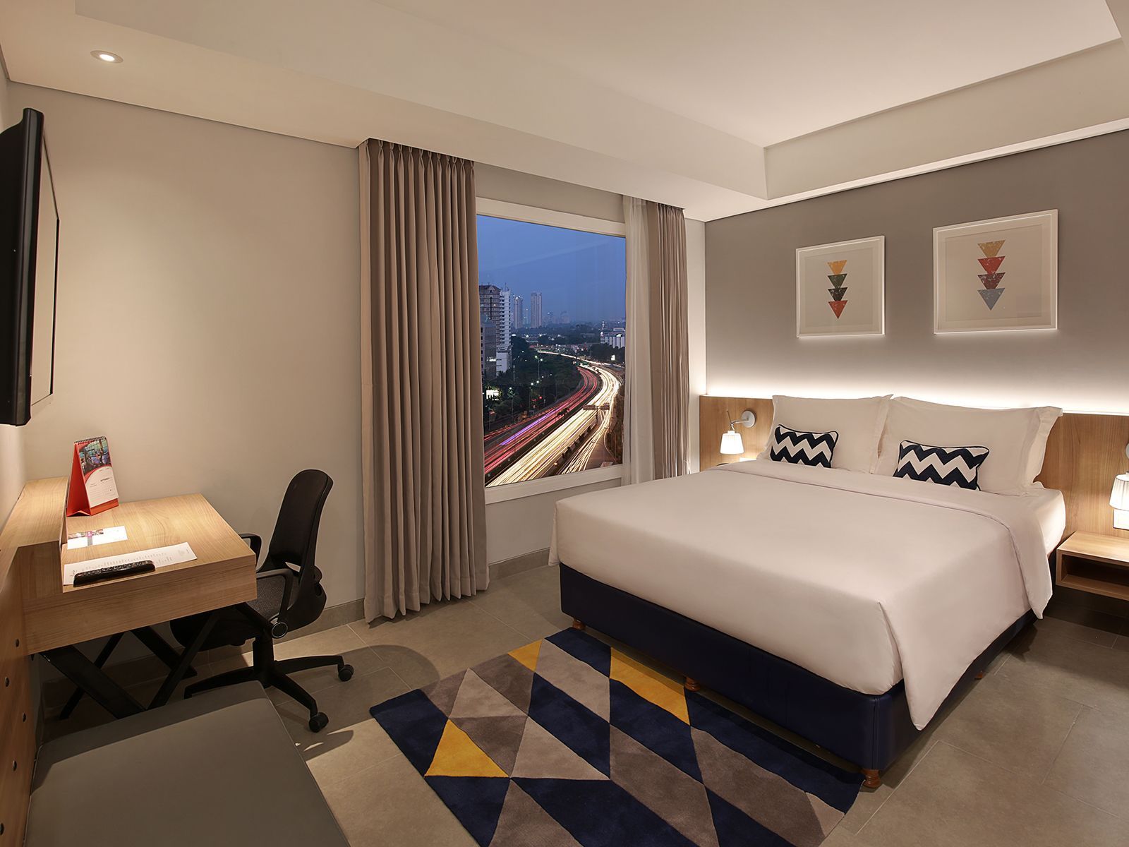 Hotel room with bed, desk, city view through window. Gray, white, and wood tones. Colorful rug.