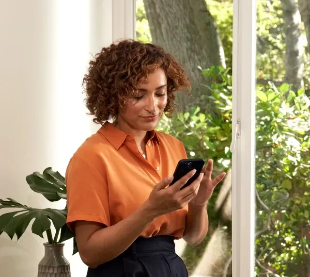 A woman in an orange shirt is looking at her cell phone setting up a sonos speaker