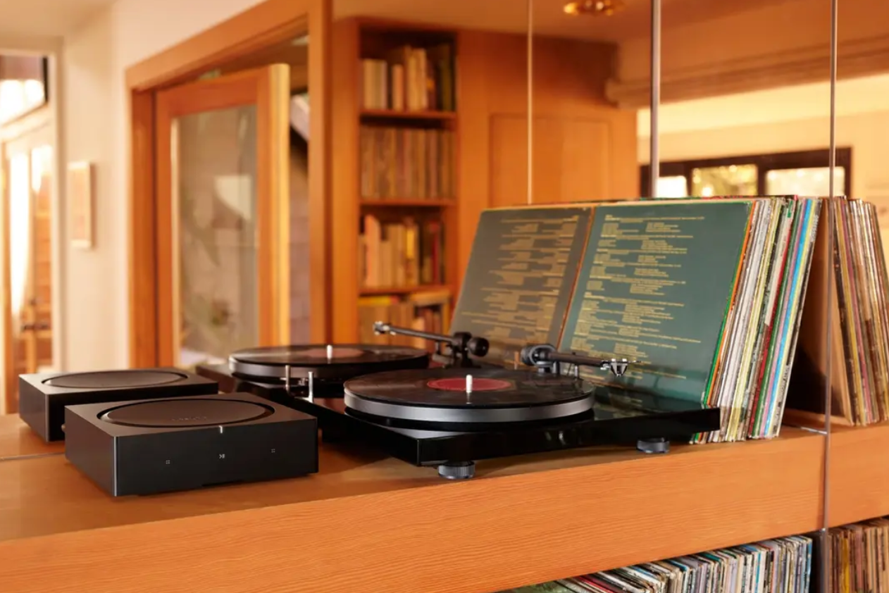 a record player is sitting on a wooden shelf next to a stack of records with a sonos amp connected to the record player for audio