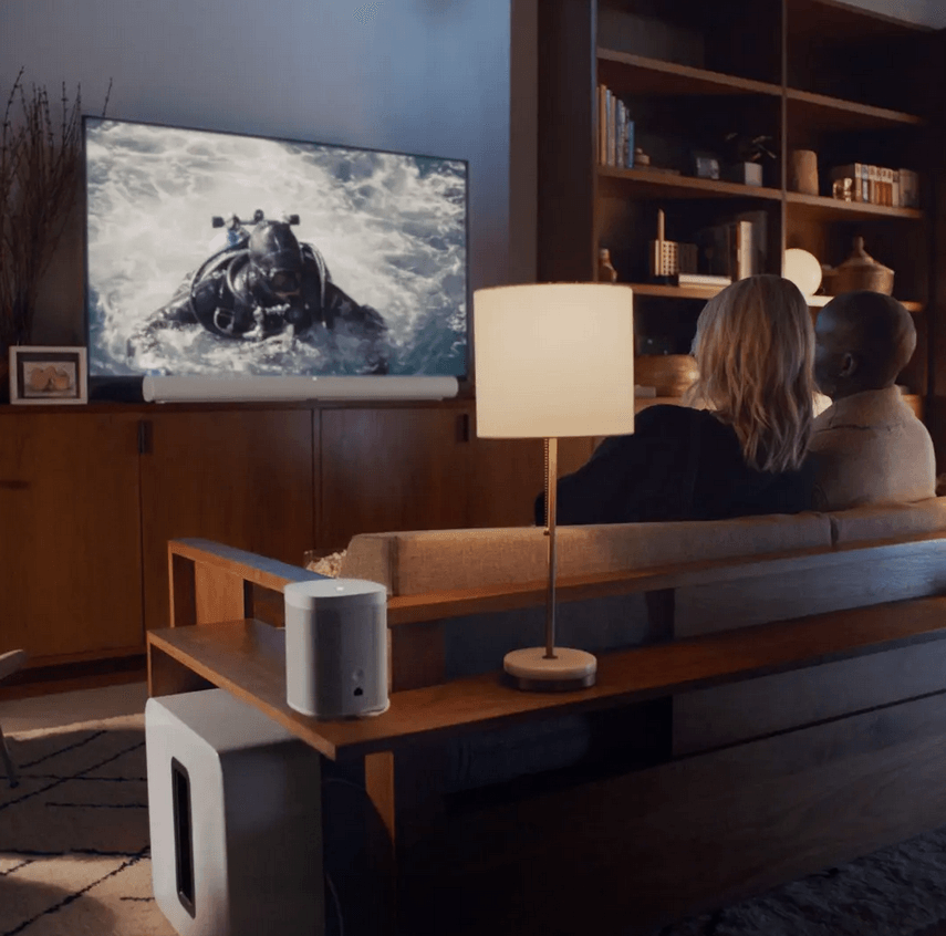 a man and woman sit on a couch watching a tv with a sonos arc soundbar while using voice services to talk to the sonos soundbar.  This arc is sold by fisher electronics in northern ohio