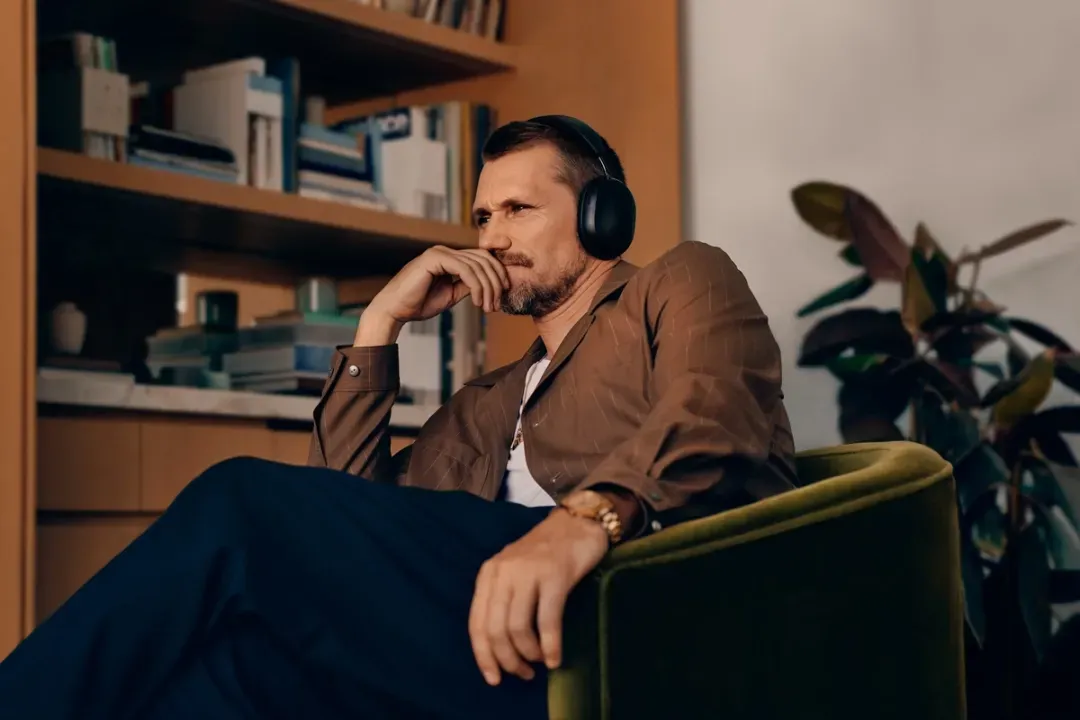 A man wearing sonos headphones is sitting in a chair in front of a bookshelf.