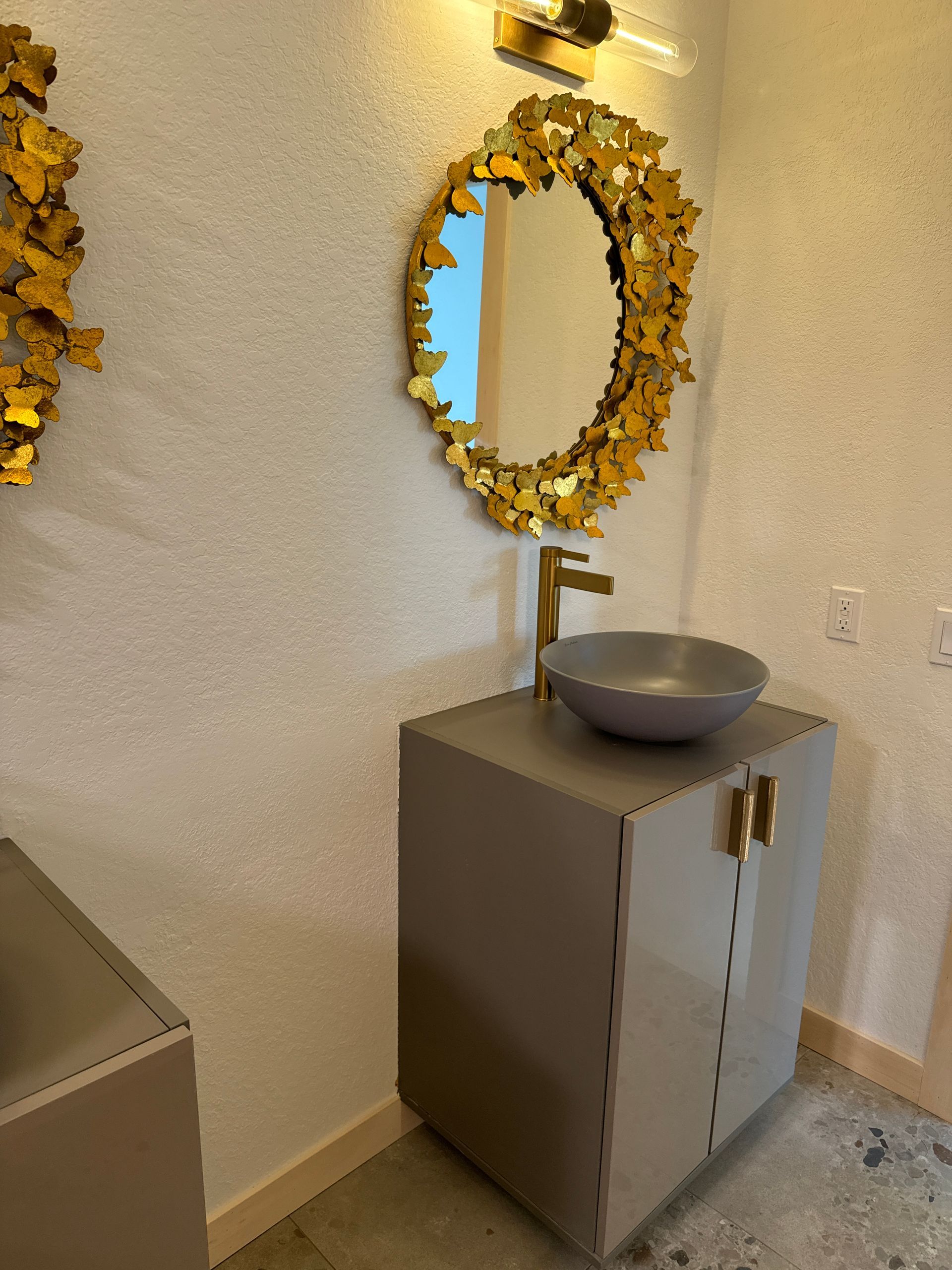 Bathroom with gray vanity, gold accents, round mirror with gold leaves, gray sink, gold faucet.