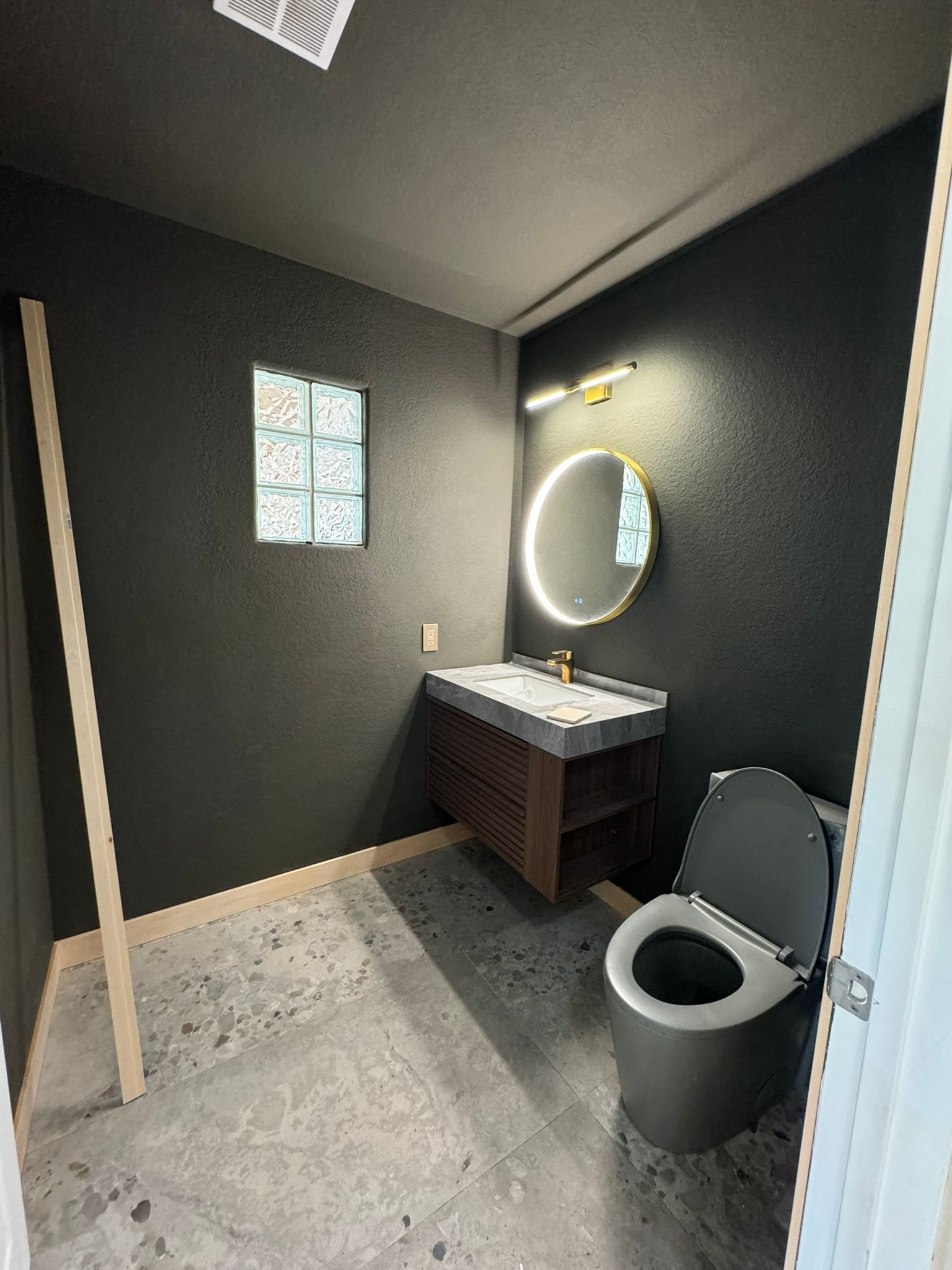 Dark-walled bathroom with a toilet, vanity, and round mirror. Concrete floor and frosted glass block window.
