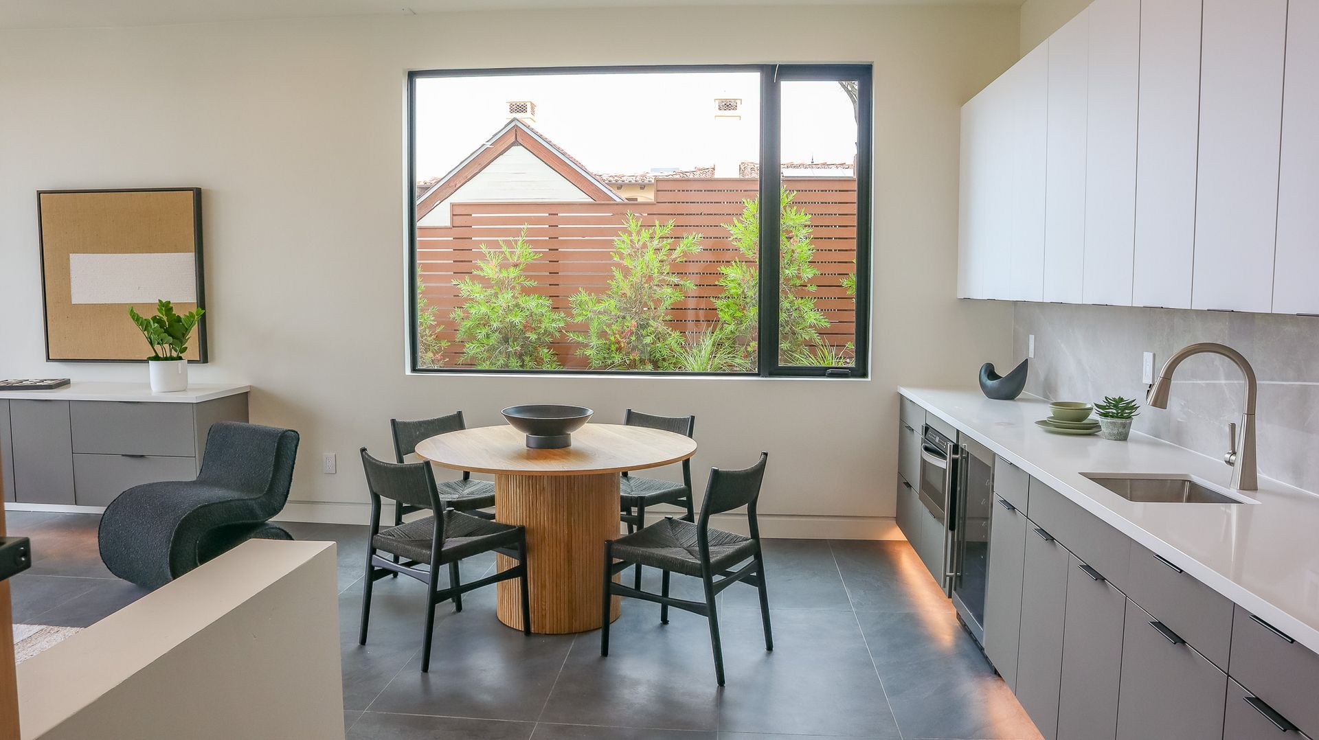 Modern kitchen and dining area with a round table, gray cabinets, and a large window overlooking a backyard.