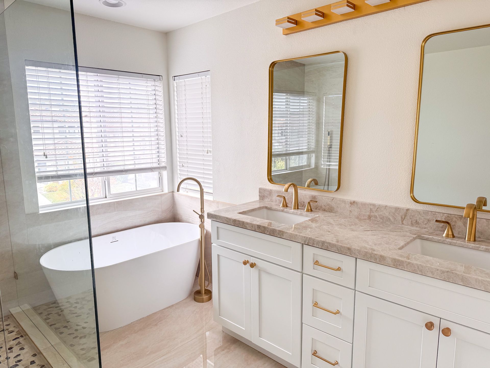 Modern white bathroom with a tub, double vanity with gold fixtures, and a window.