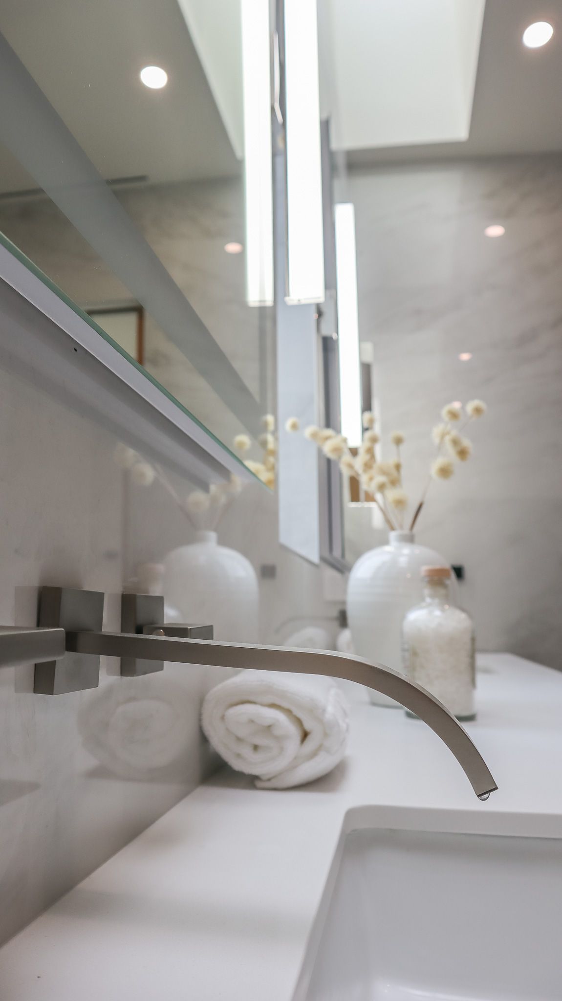 Bathroom sink with faucet, rolled towels, white vases, and tall narrow lights reflecting in a mirror.
